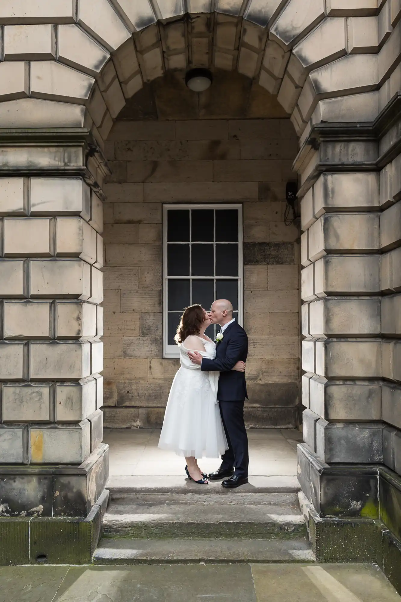 A couple in formal attire embrace and share a kiss under a stone archway of a historic building.