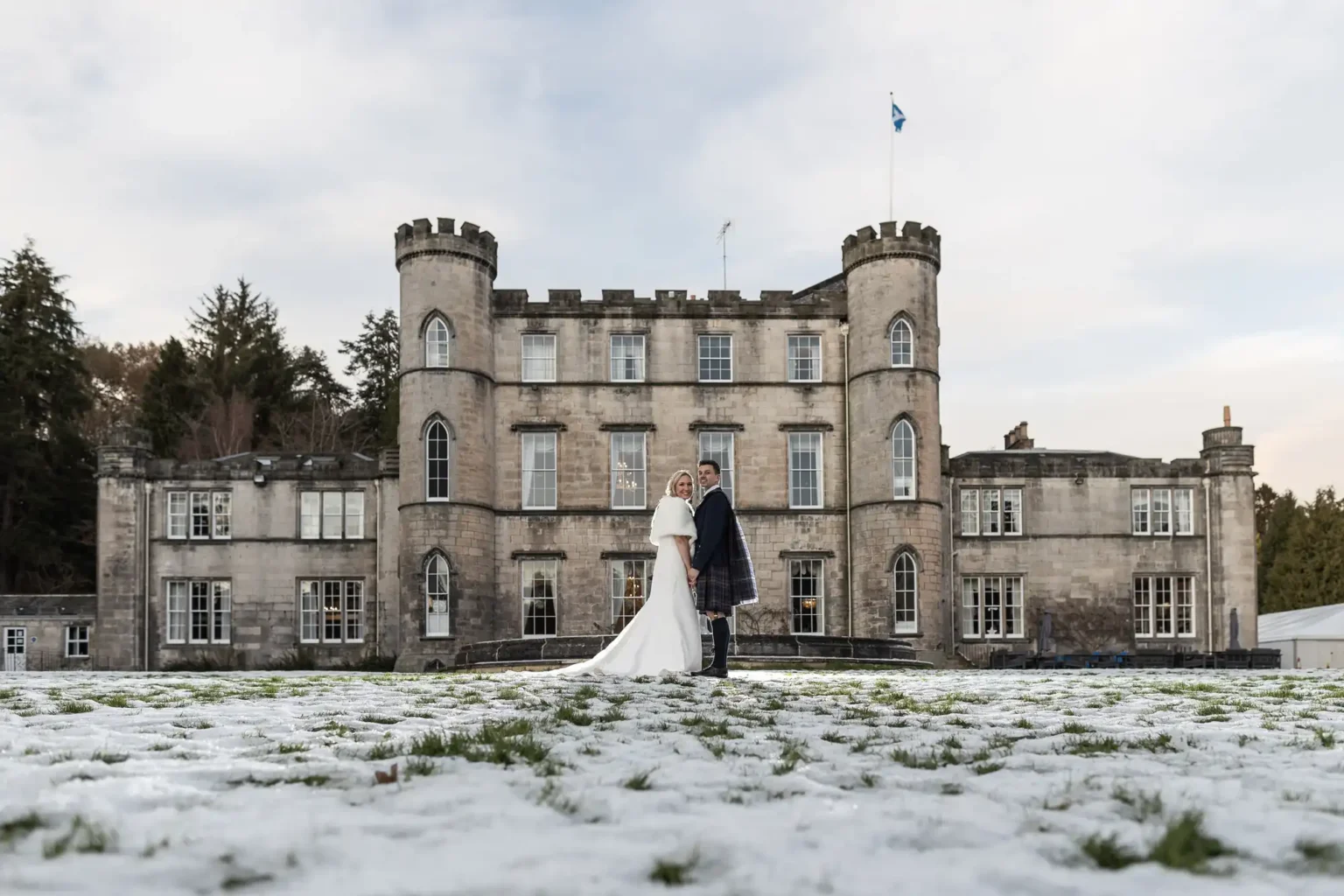 A bride and groom stand together on snow-dusted grass in front of a large stone castle with round towers.