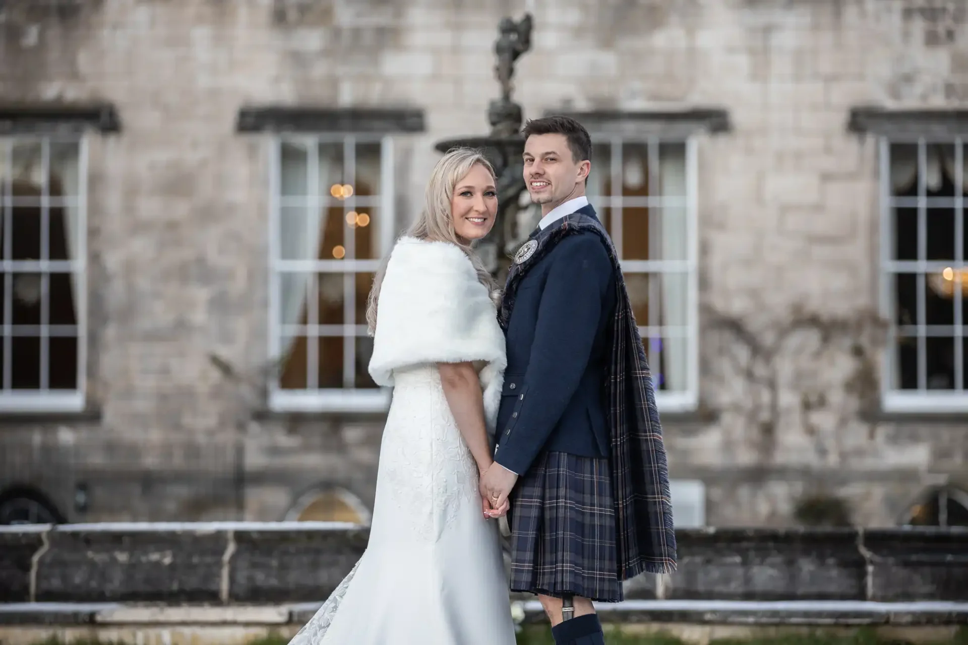 A bride in a white dress and a groom in a kilt stand holding hands outdoors in front of a stone building, both smiling at the camera.