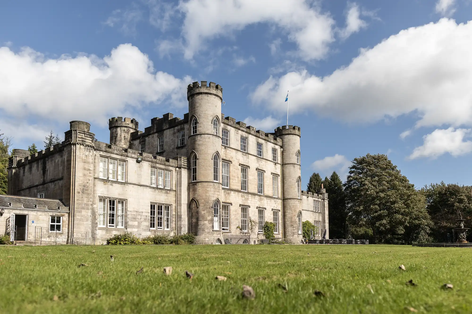 A large stone castle with turrets and battlements stands under a partly cloudy sky, surrounded by an expanse of green grass and trees.