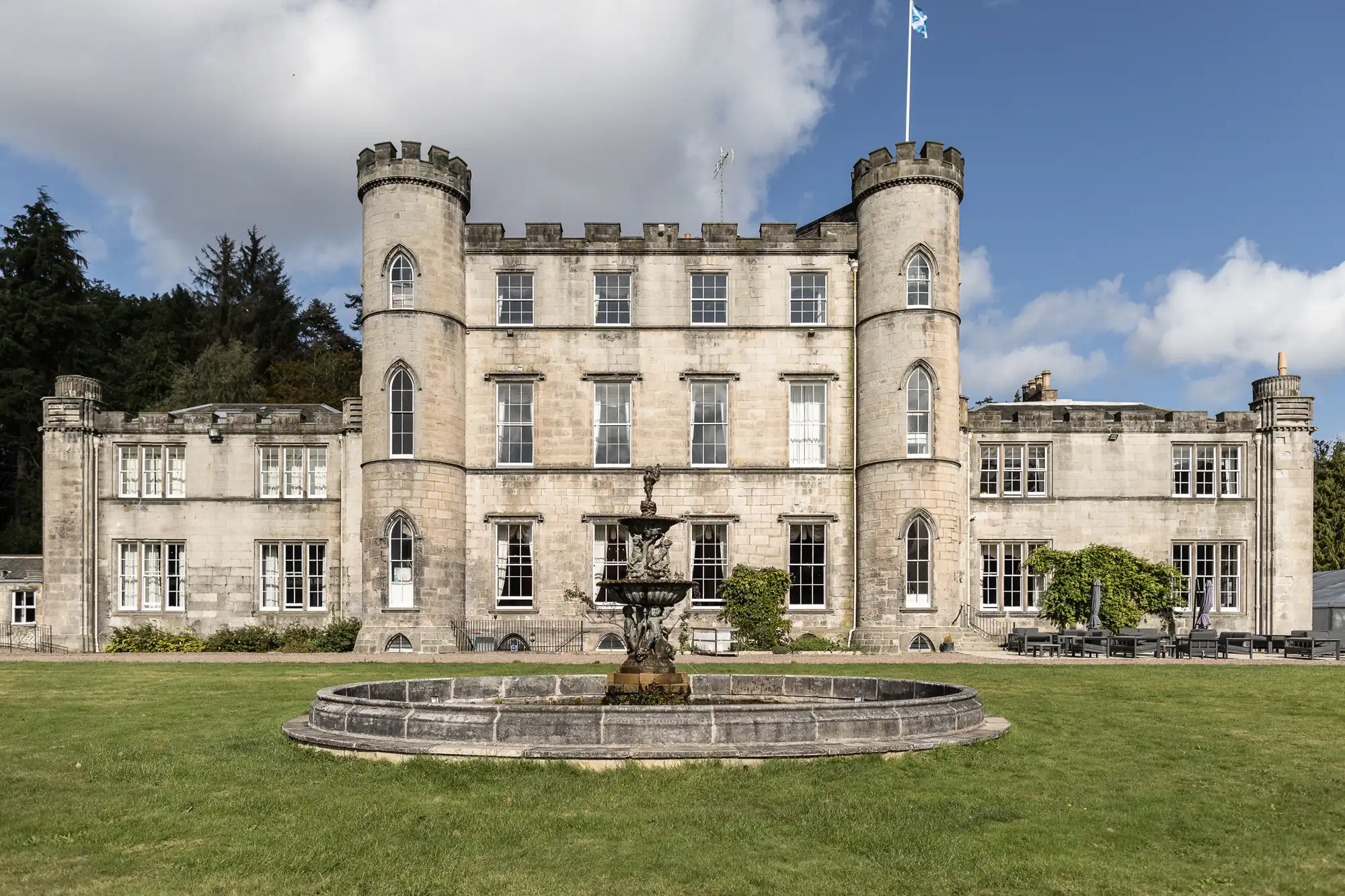 A large stone castle with two round towers, multiple windows, and a central fountain in front, set against a backdrop of trees and a blue sky with some clouds.