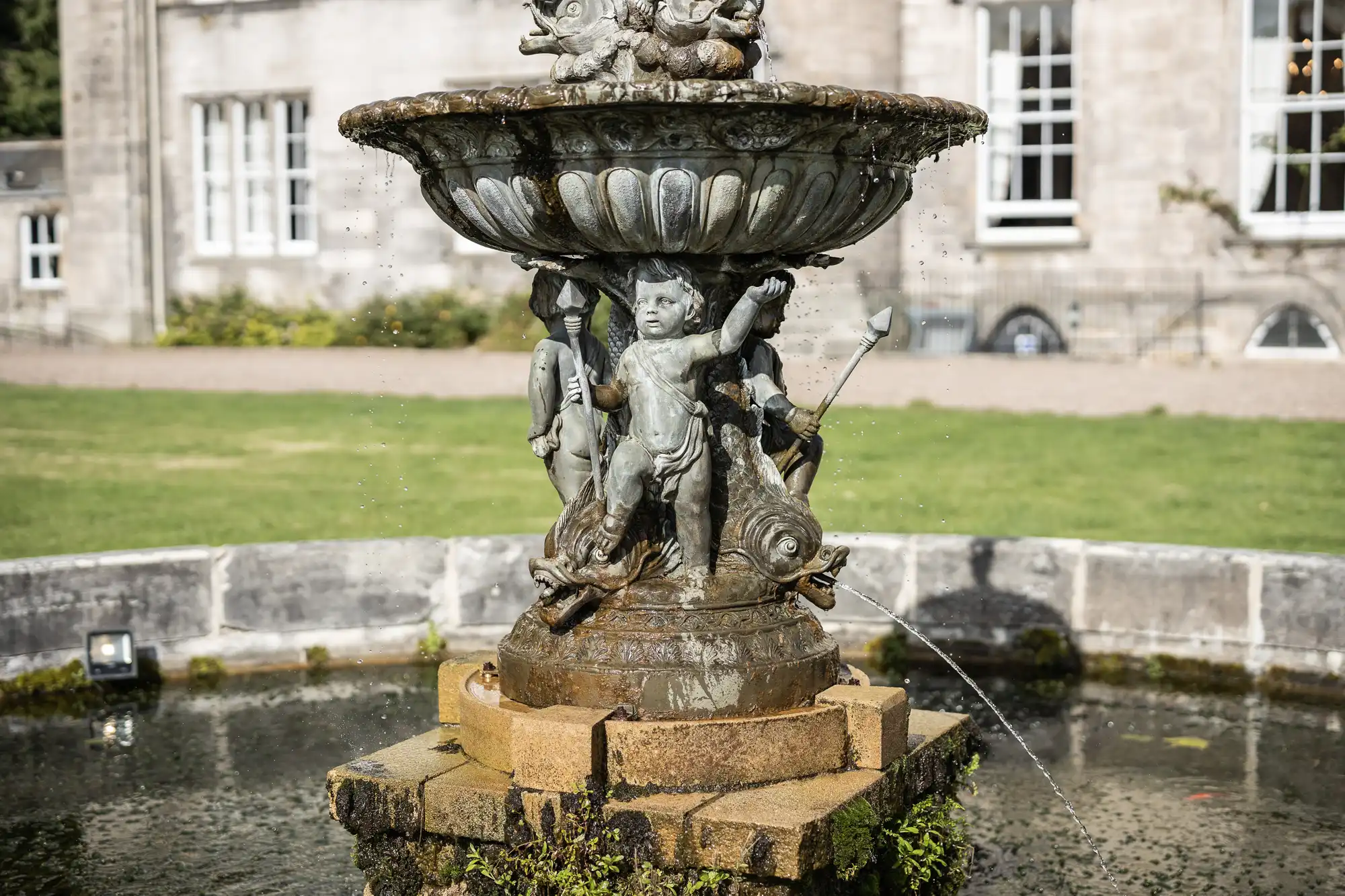 A decorative stone fountain features cherubic figures and sea creatures. It is situated in a grassy area near a building. Water streams from the mouths of the sea creatures into the basin below.