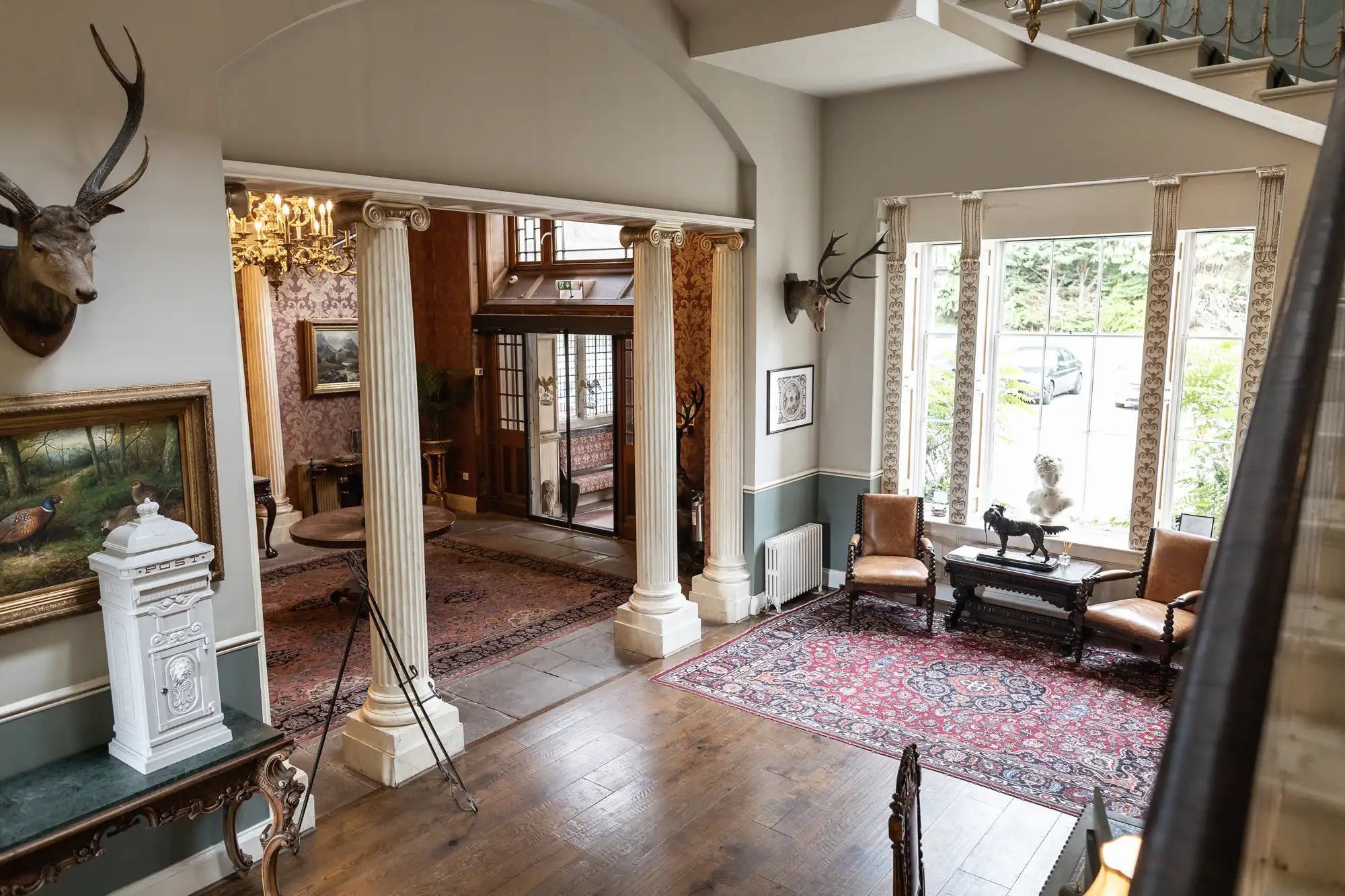 A spacious interior with an archway, columns, a large window, wooden floors, area rugs, and antique furniture. Deer head and painting adorn the walls. An entryway can be seen in the background.