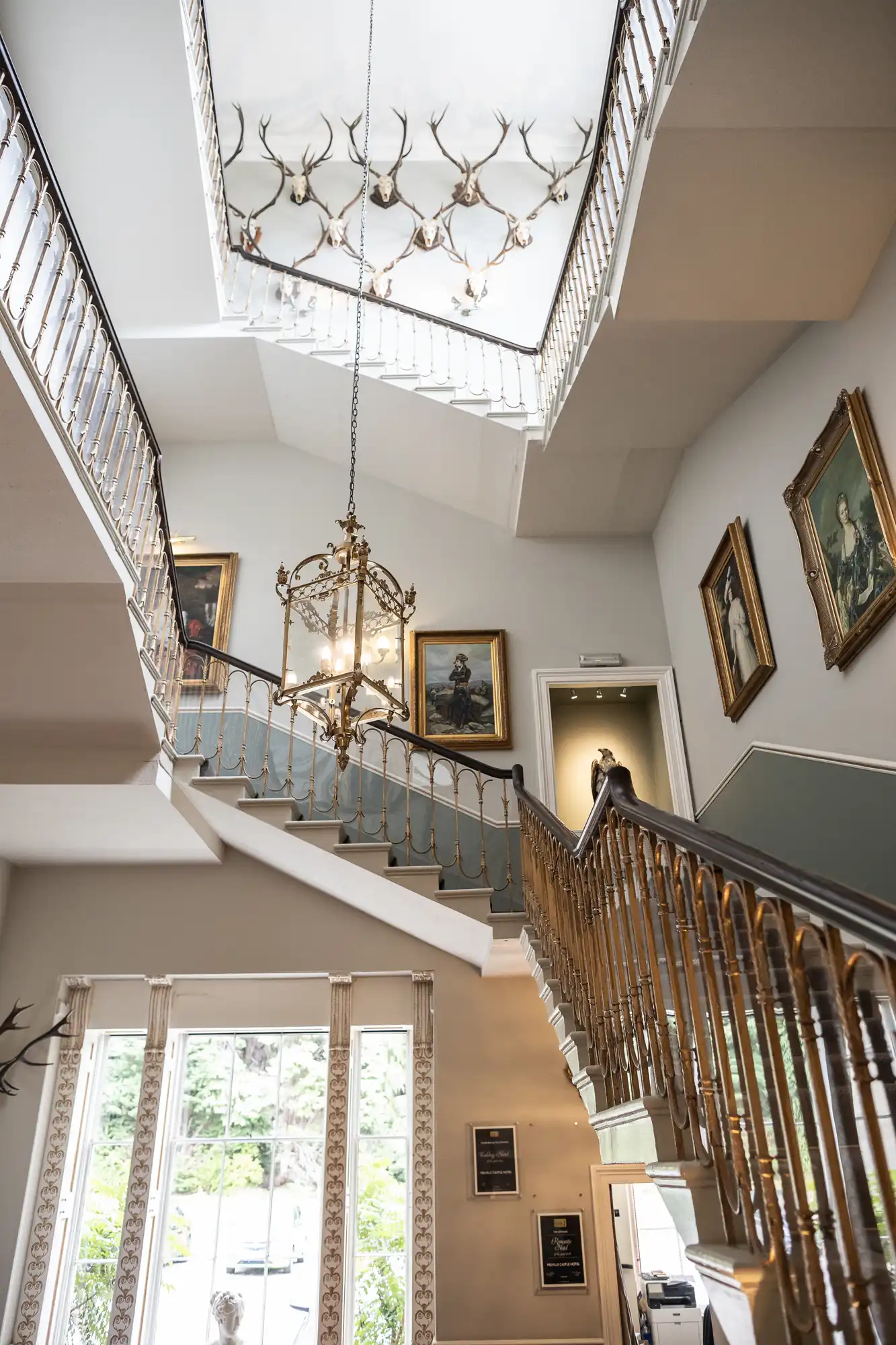 Grand staircase with ornate brass railings, chandelier, and numerous antlers and framed portraits adorning the walls. Large windows allow natural light to illuminate the space.