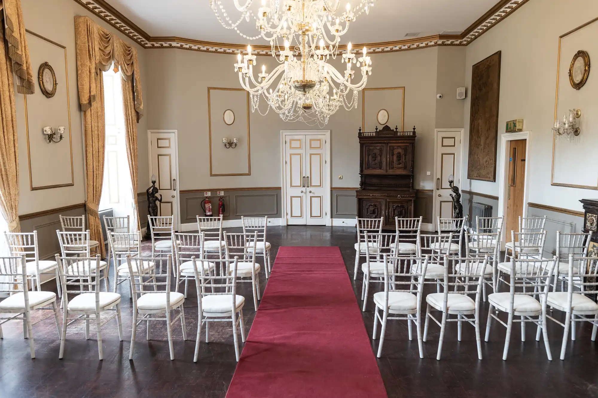 A formal event room with white chairs arranged in rows facing a central aisle covered with a red carpet. Chandeliers hang from the ceiling, and the room features ornate golden decor and large curtains.