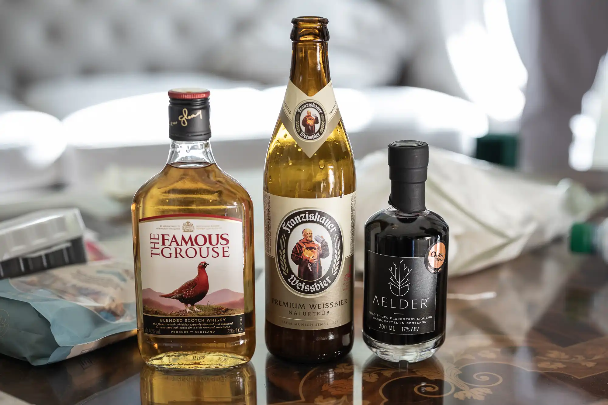 Three bottles displayed on a table: a bottle of The Famous Grouse Scotch whisky, a bottle of Franziskaner Weissbier, and a bottle of Aelder Elixir. Items and packaging are visible in the background.