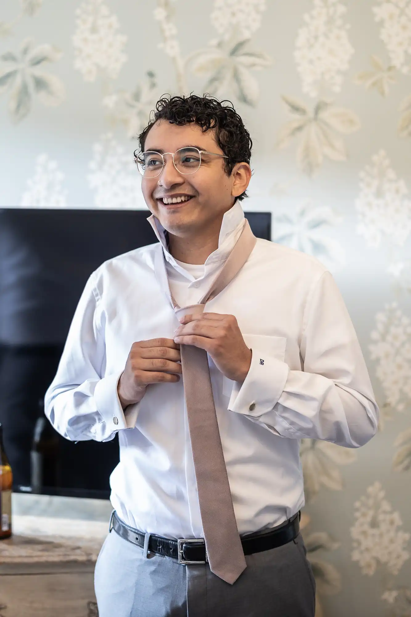 A man with curly hair and glasses is smiling while putting on a beige tie in a room with floral wallpaper.