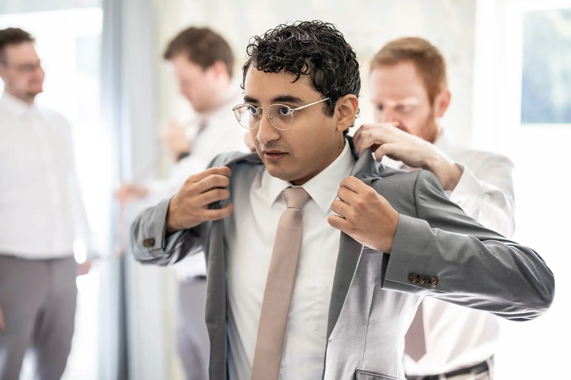 A man with glasses and a tie adjusts his gray suit jacket, assisted by another man. Two other men in white shirts are visible in the background.