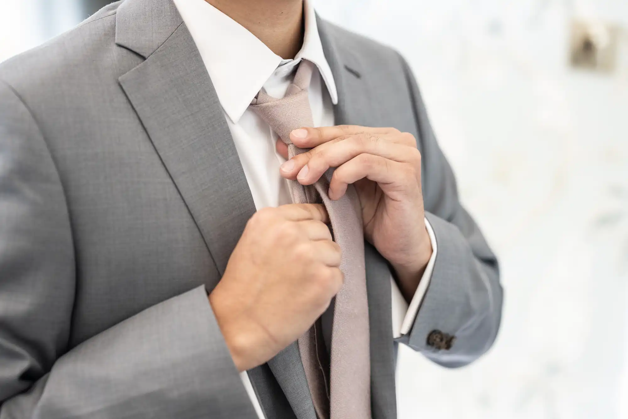 Person in a gray suit adjusting their light pink tie with both hands, while wearing a white dress shirt.