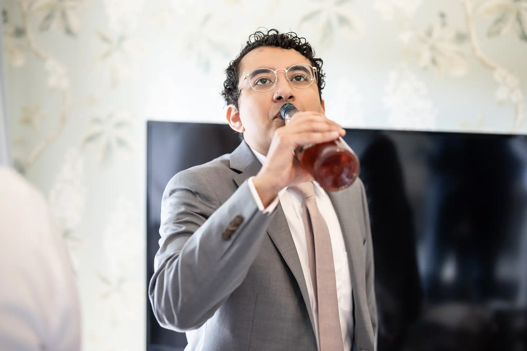 A man in a gray suit and glasses is drinking from a bottle. A television screen is visible in the background.