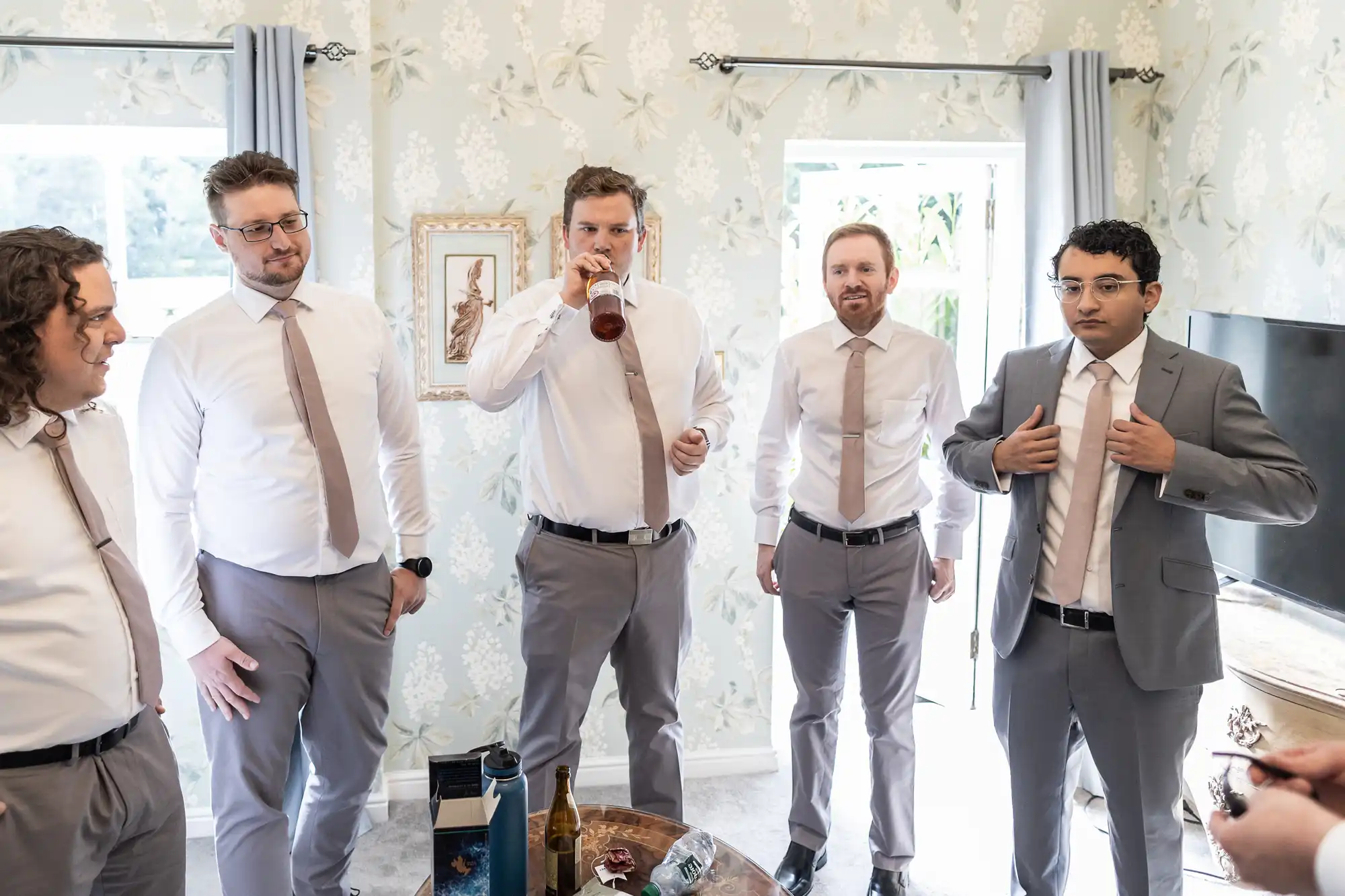 Five men in light gray suits and beige ties are standing in a room decorated with floral wallpaper; one is drinking from a bottle, and another is adjusting his jacket. Bottles and snacks are on the table.