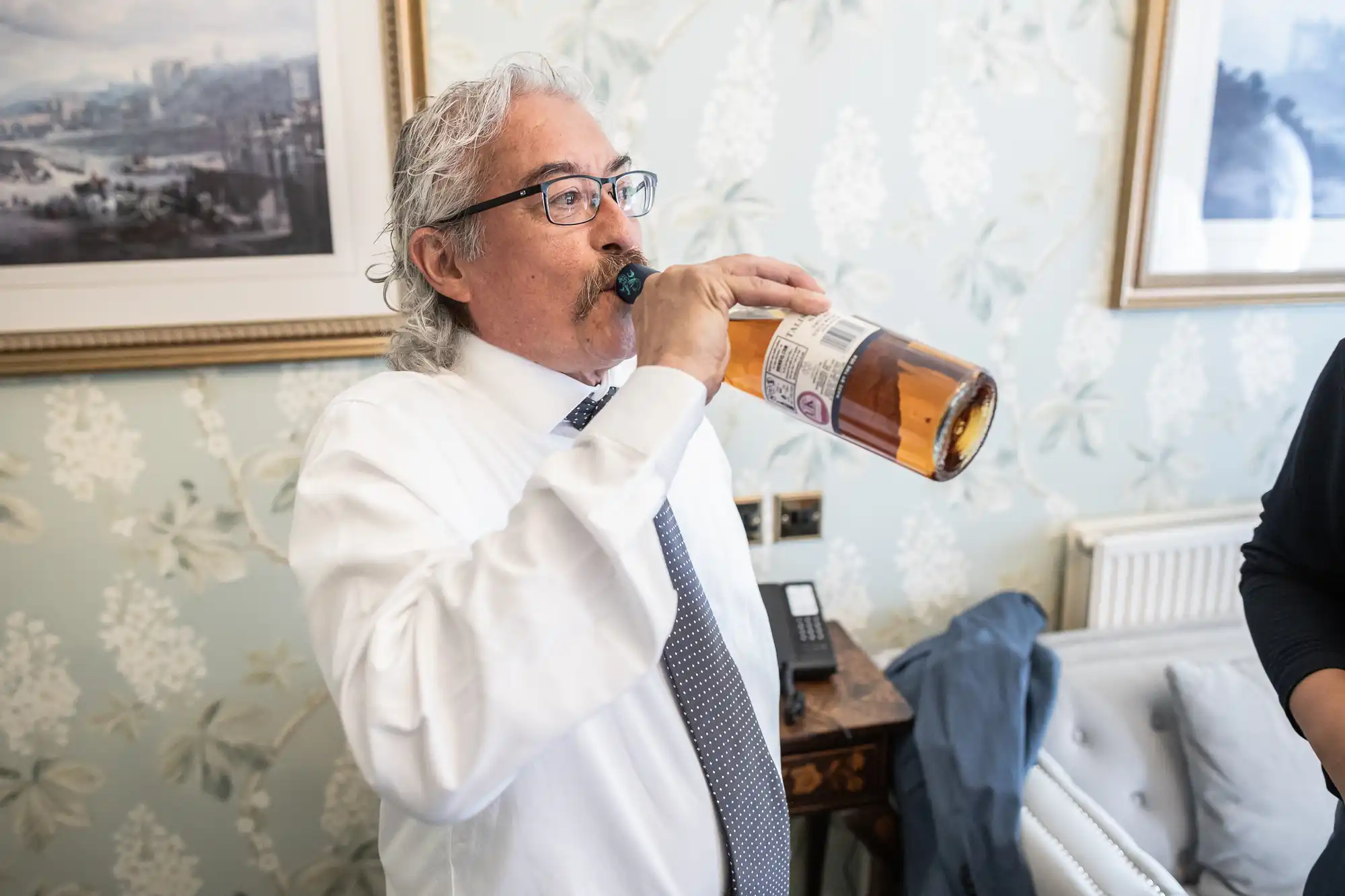 A man with grey hair and glasses, dressed in a white shirt and tie, drinks directly from a bottle in a room with floral wallpaper and framed pictures.