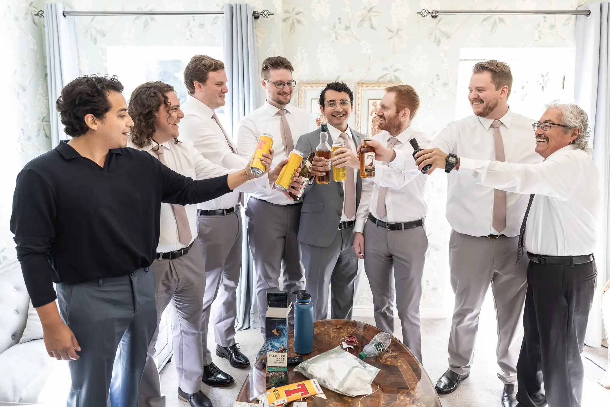 A group of men, some in suits and some in casual attire, gather in a well-lit room, holding drinks and toasting each other. They seem to be celebrating or preparing for an event.