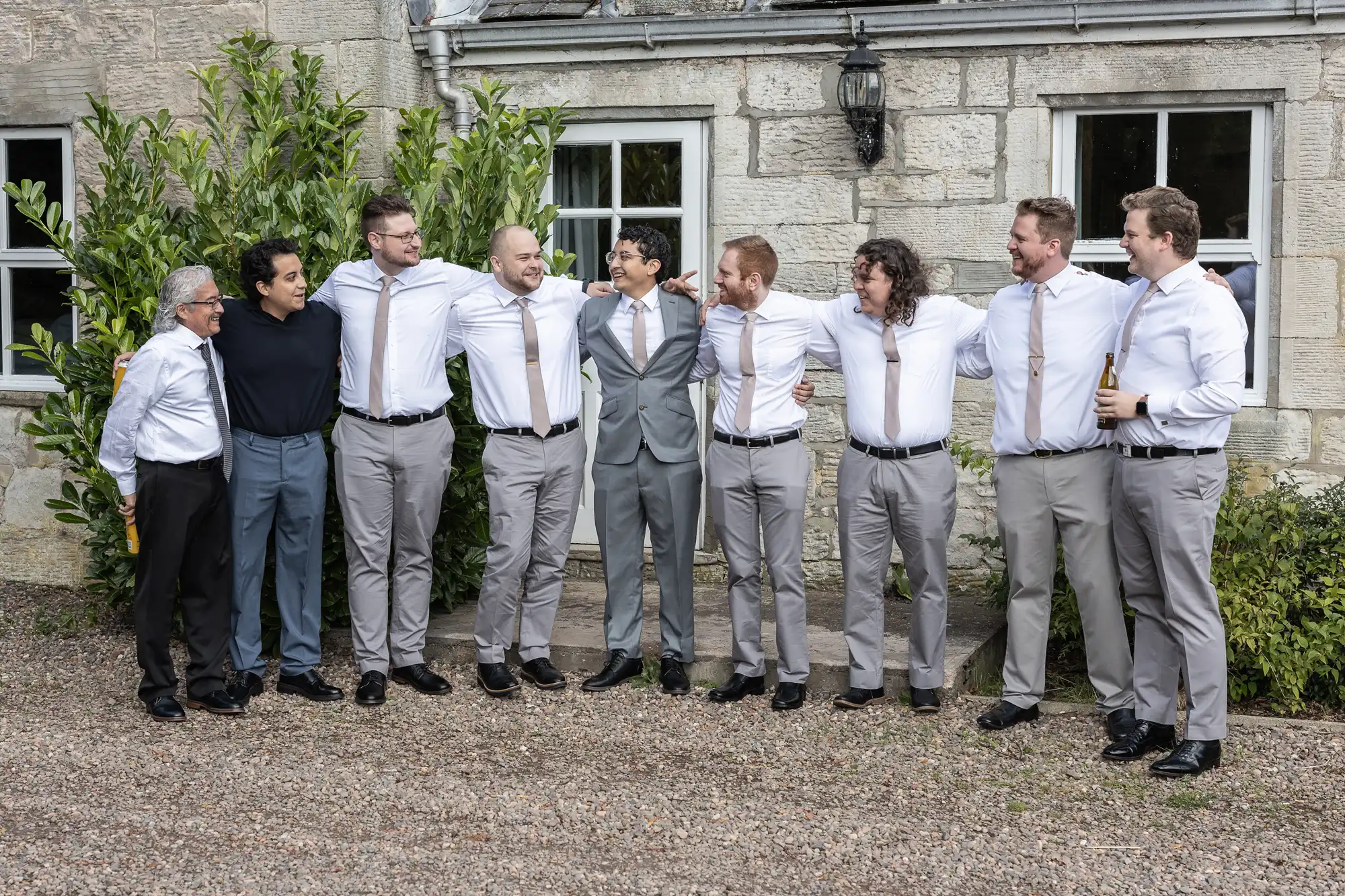 A group of eight men, most dressed in white shirts and light grey pants, stand arm in arm outside a stone building. One man wears a grey suit and another a black shirt.