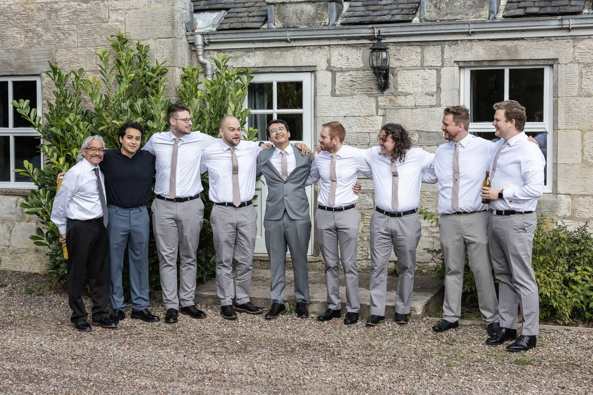 A group of nine people, dressed in formal wear, stand in a line with their arms around each other in front of a stone building with windows and plants.