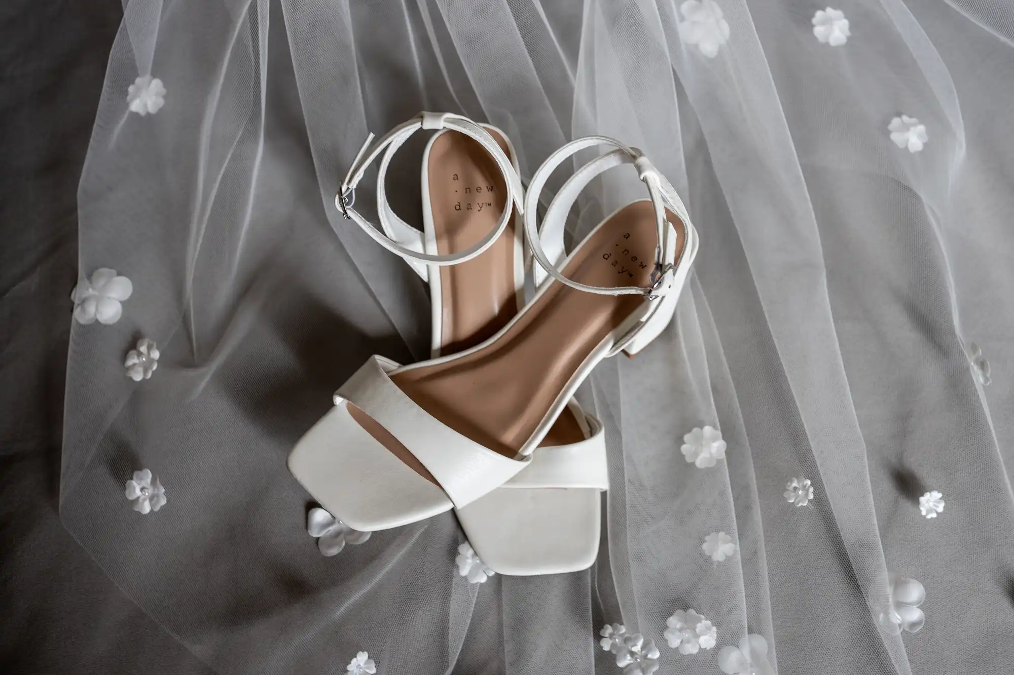 White bridal shoes with ankle straps placed on a gray, flower-embroidered veil.