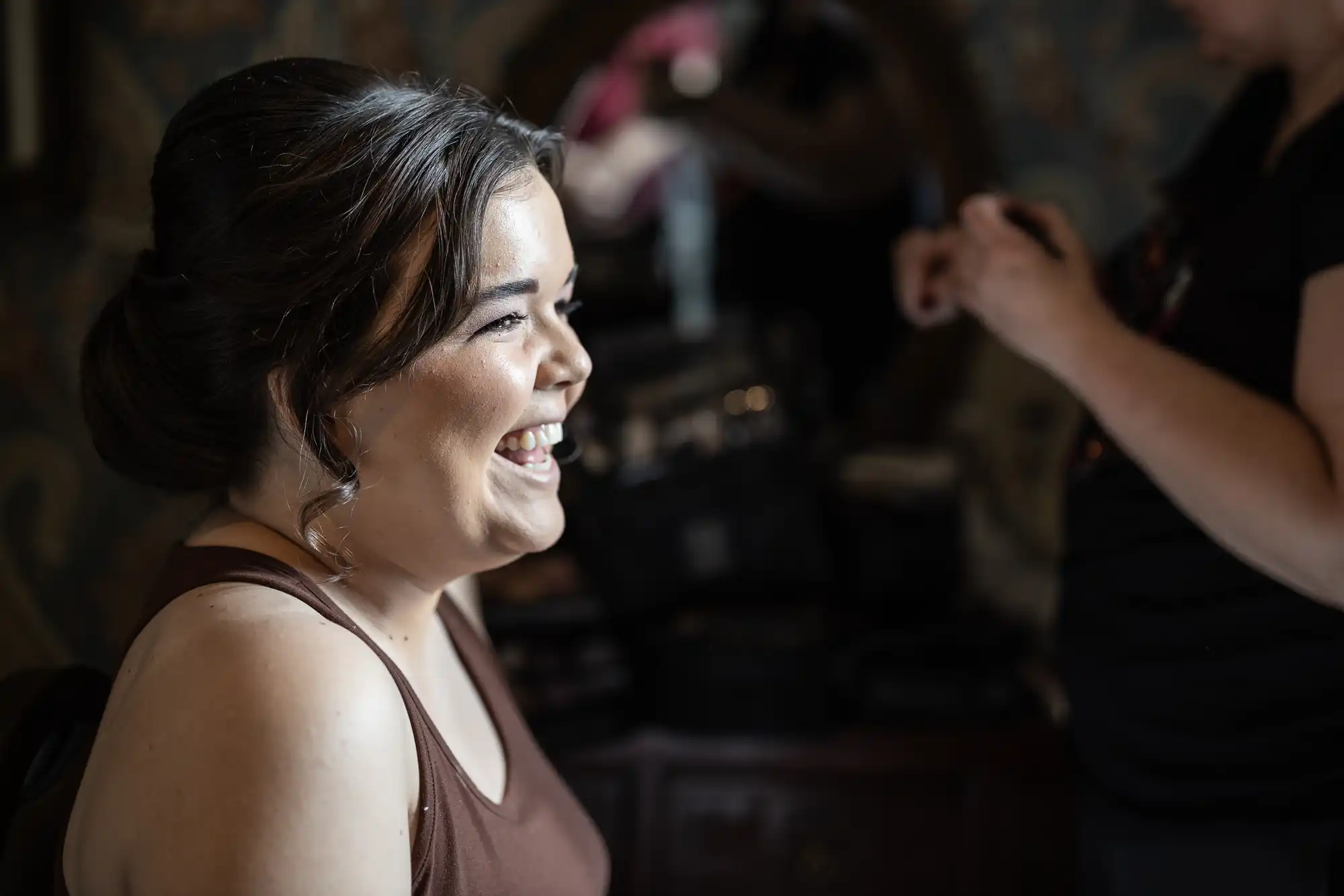 A woman with dark hair, wearing a brown top, smiles while sitting indoors. Another person is in the background, partially visible, holding some items.