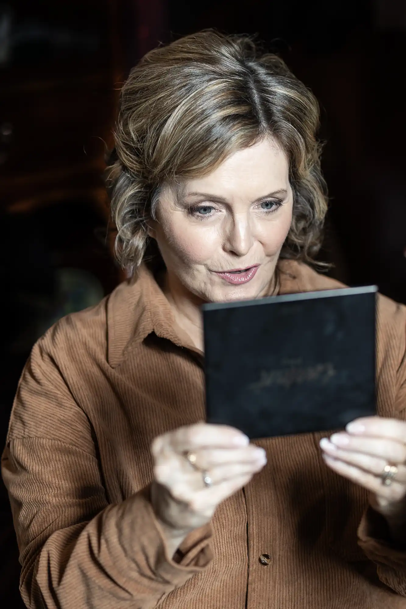 A woman with short, curly hair wearing a brown jacket looks at a small black rectangular object she is holding.