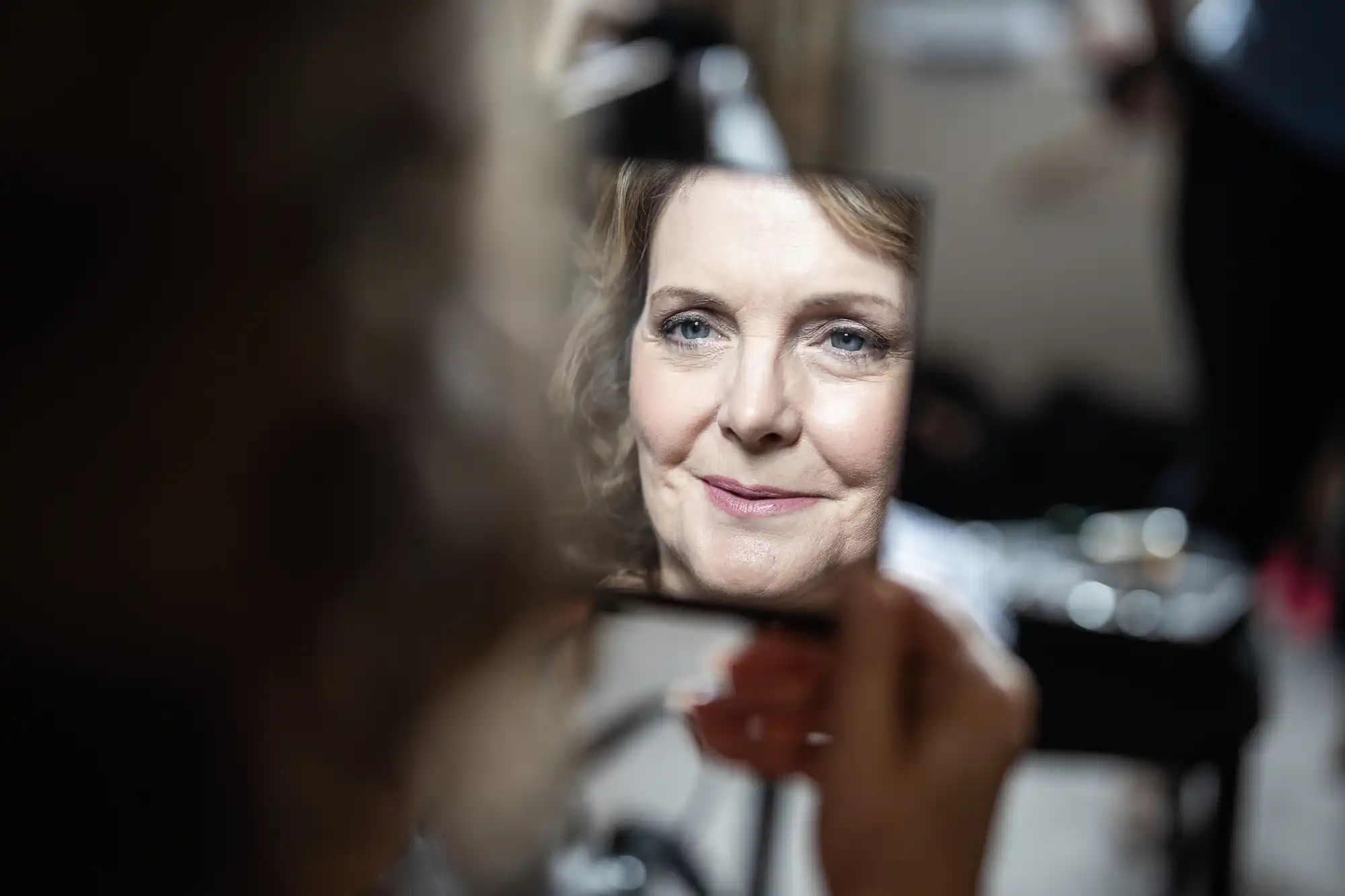 A woman looks at her reflection in a handheld mirror, which she holds close to her face.