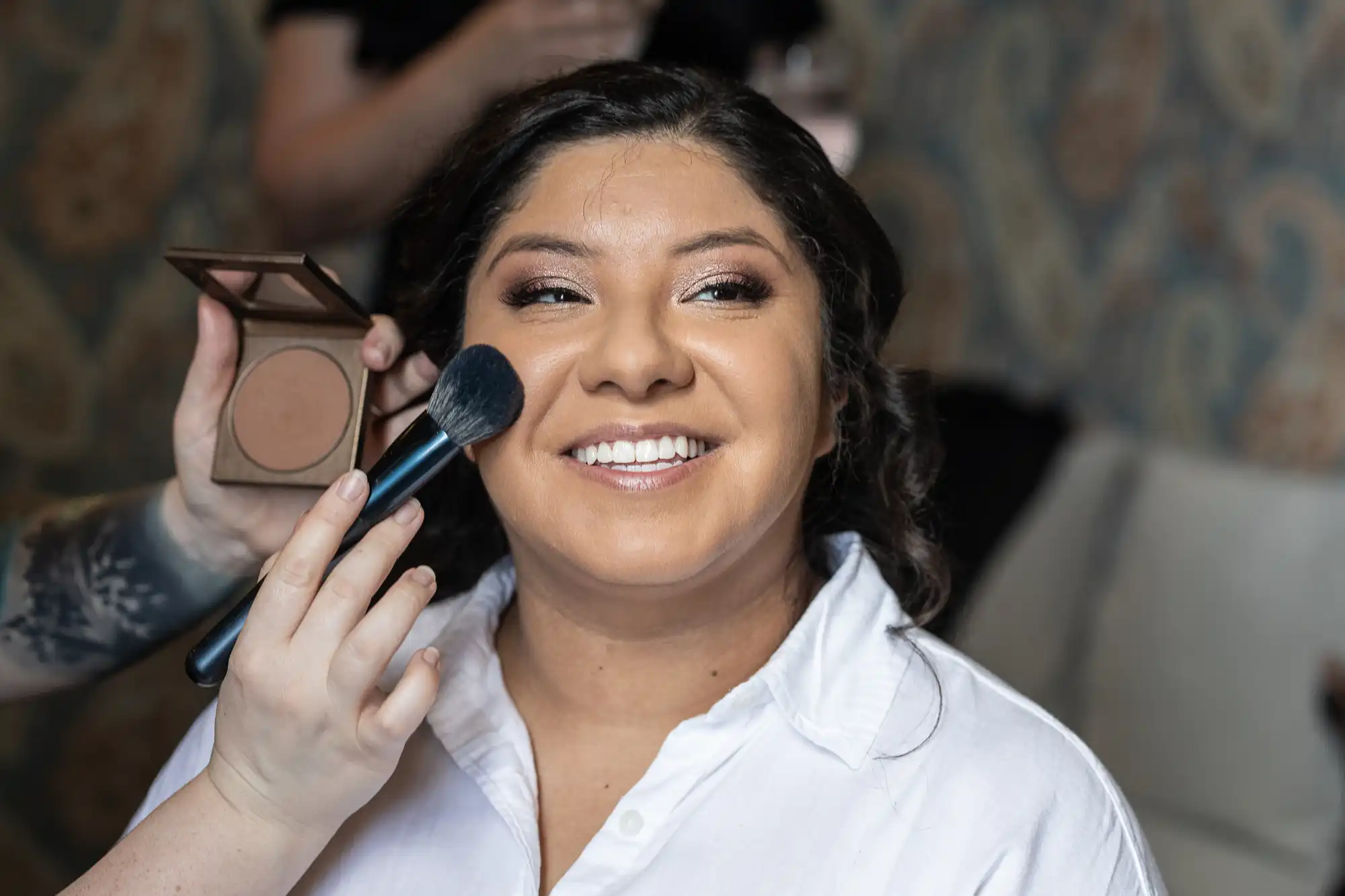 A woman is smiling while having makeup applied to her face with a brush. She is wearing a white shirt and sitting indoors.