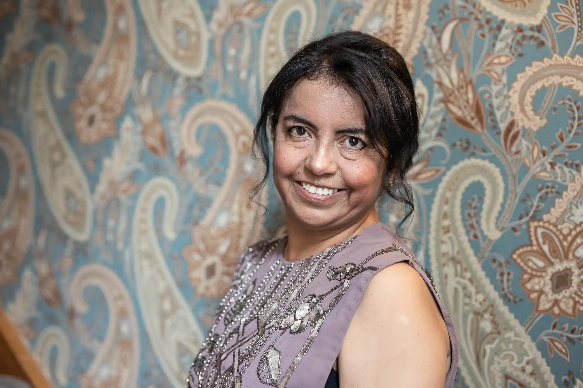 A woman with dark hair smiles while standing in front of a detailed, paisley-patterned wallpaper. She is wearing a sleeveless top adorned with sequins.