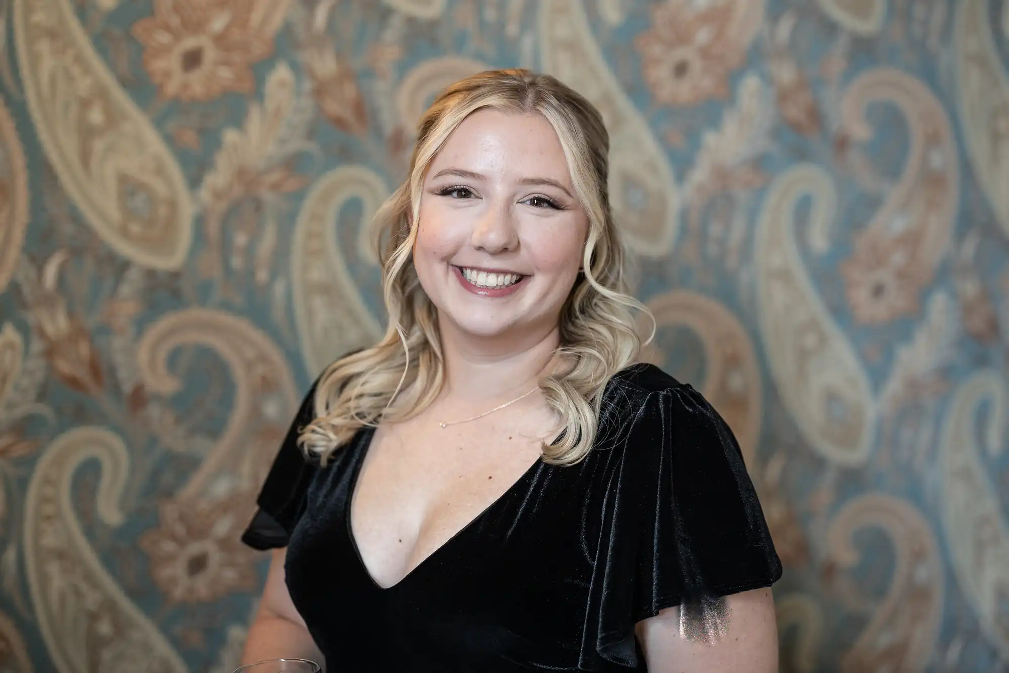 A woman with blonde hair wearing a black dress smiles at the camera against a patterned background.