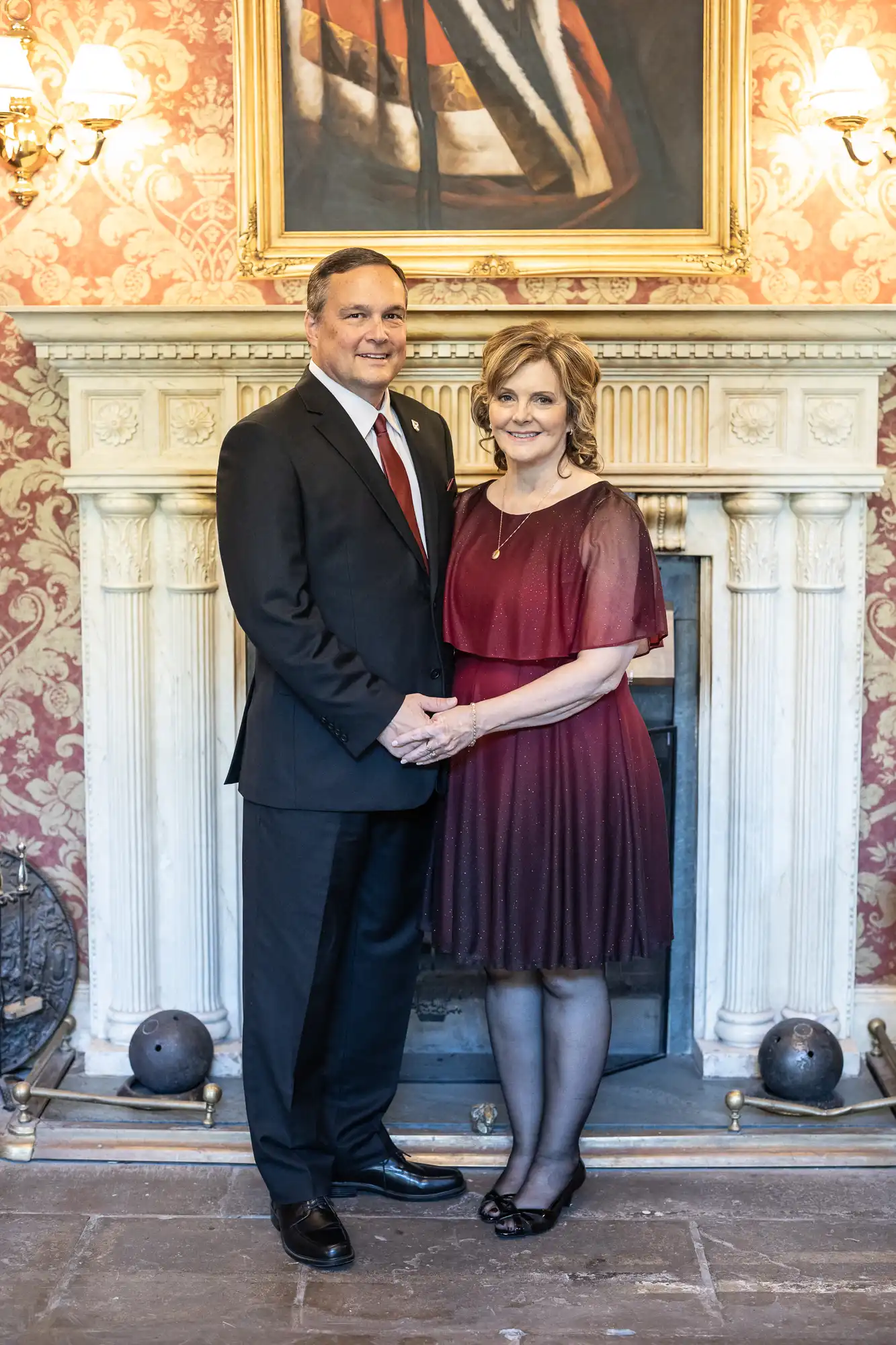 A man and a woman stand side by side holding hands in front of a fireplace. Both are dressed formally, with the man in a suit and tie and the woman in a red dress.