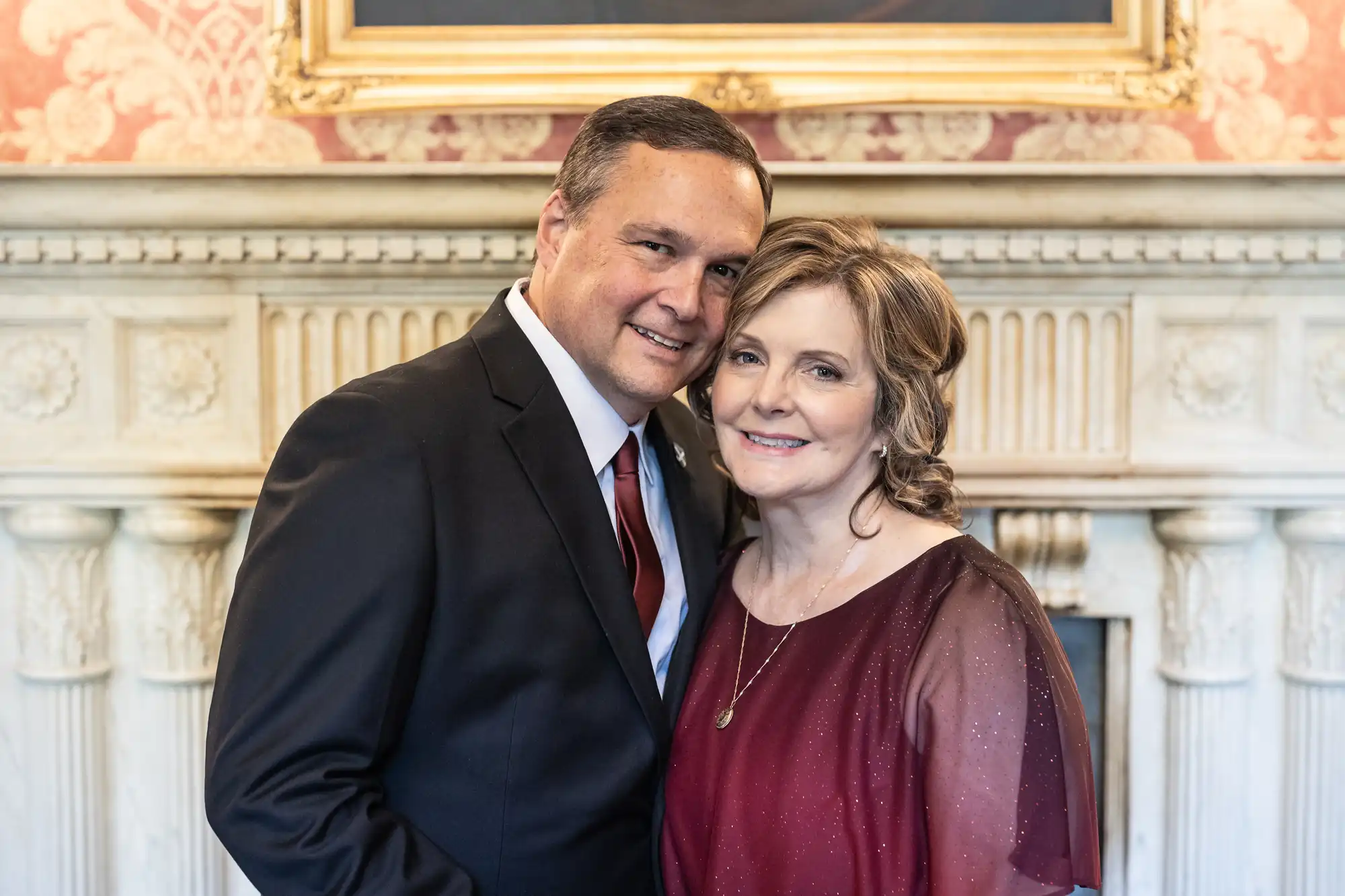 A man in a black suit and red tie stands beside a woman in a burgundy dress, both smiling in front of an ornate fireplace with a framed painting above it.