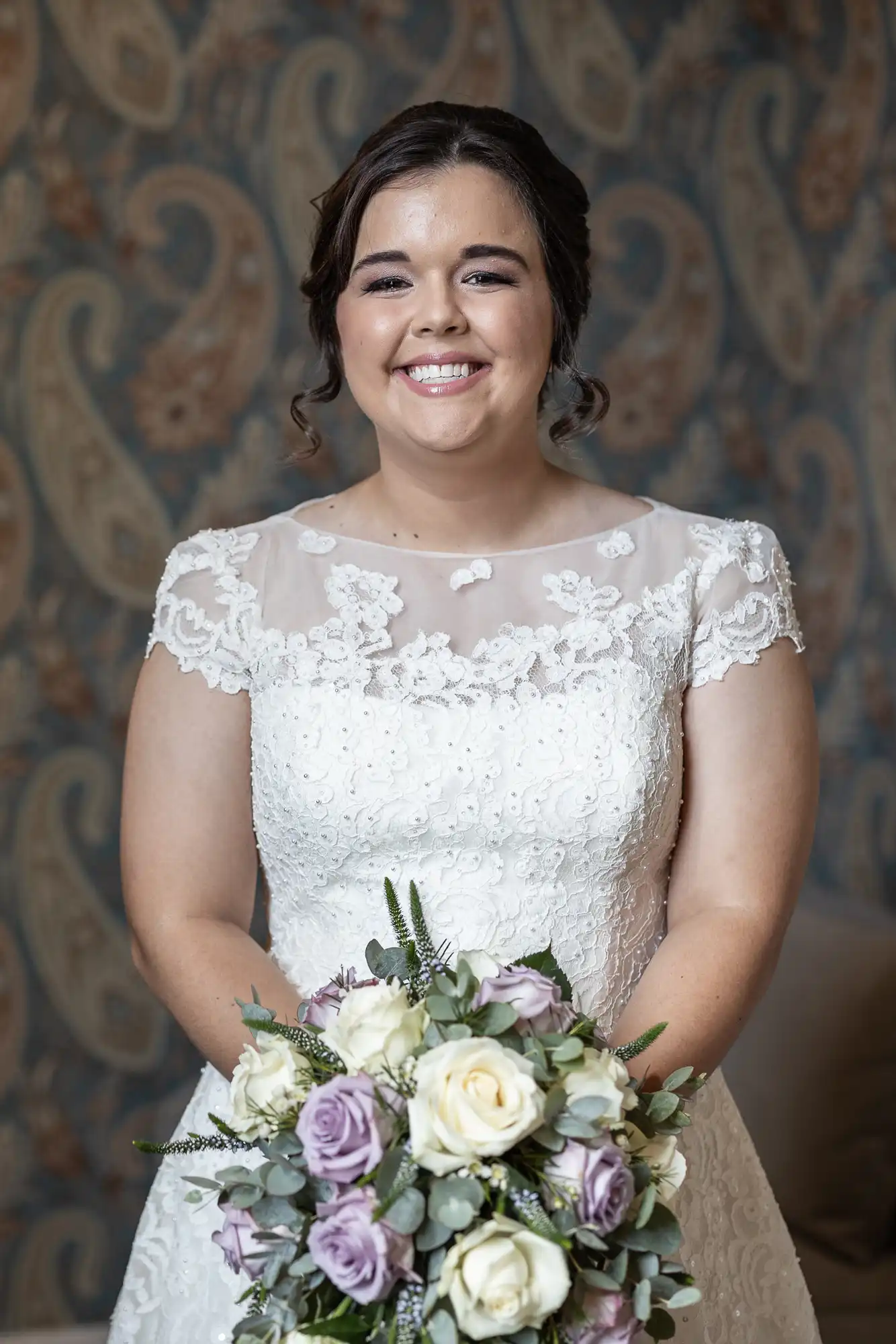 A woman in a white lace wedding dress smiles while holding a bouquet of purple and white roses, standing indoors against a patterned background.