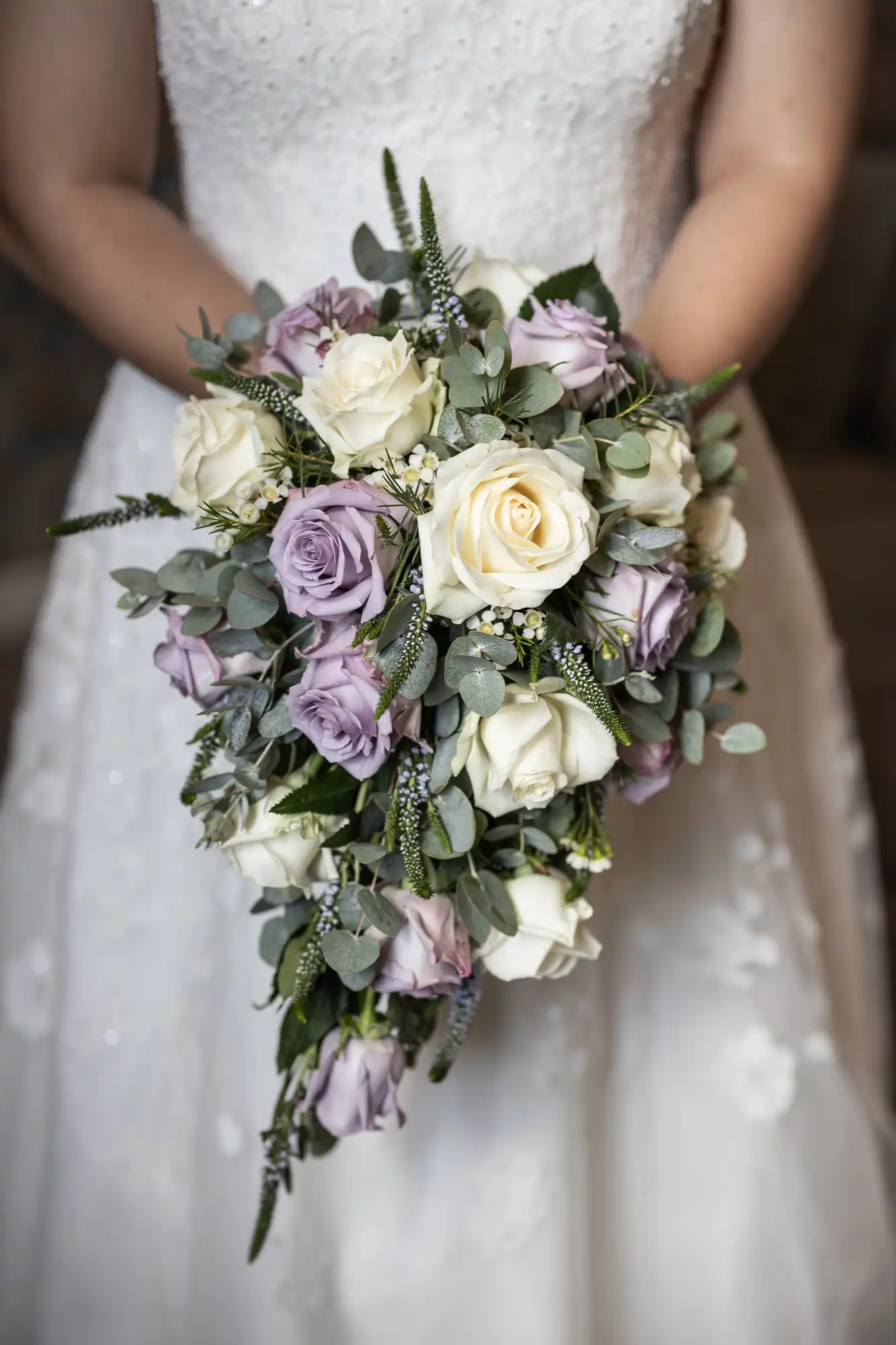 A bride in a white lace dress holds a cascading bouquet of white and purple roses mixed with greenery.