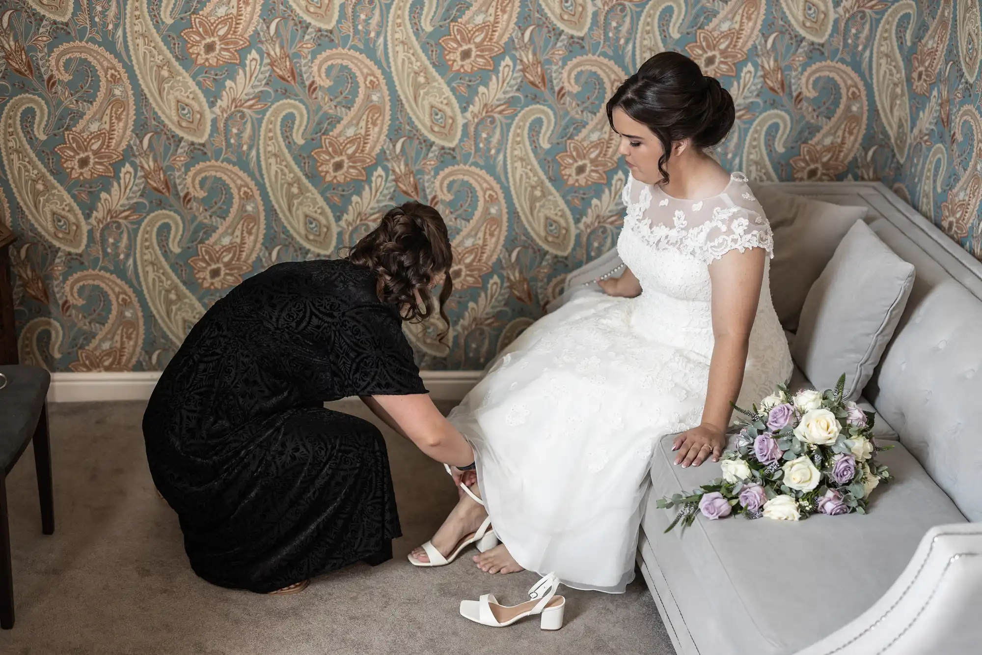 A woman in a white wedding dress sits on a couch while another woman in a black dress helps her with her shoes. A bouquet of flowers rests beside the bride.