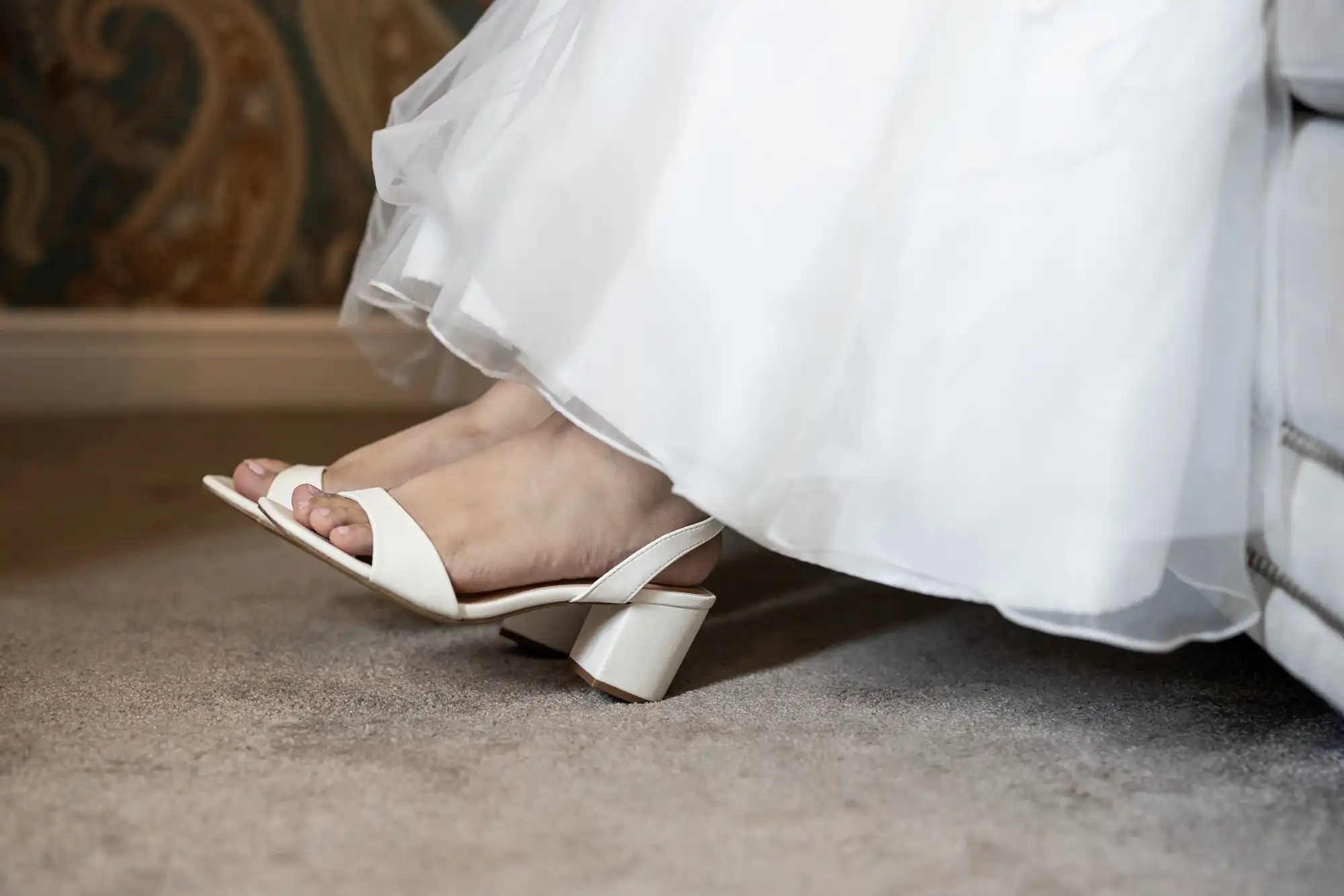 A person in a white dress wearing white, open-toed heels, with their heel lifted from a carpeted floor.