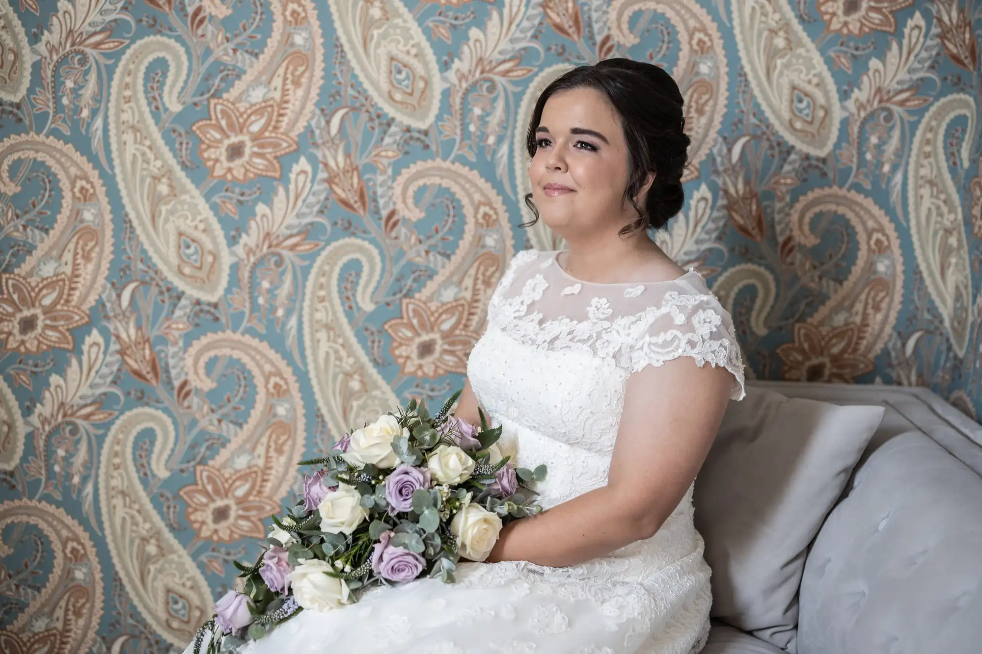 A bride in a white lace wedding dress holds a bouquet of white and purple flowers while sitting against a patterned, floral wallpaper backdrop.