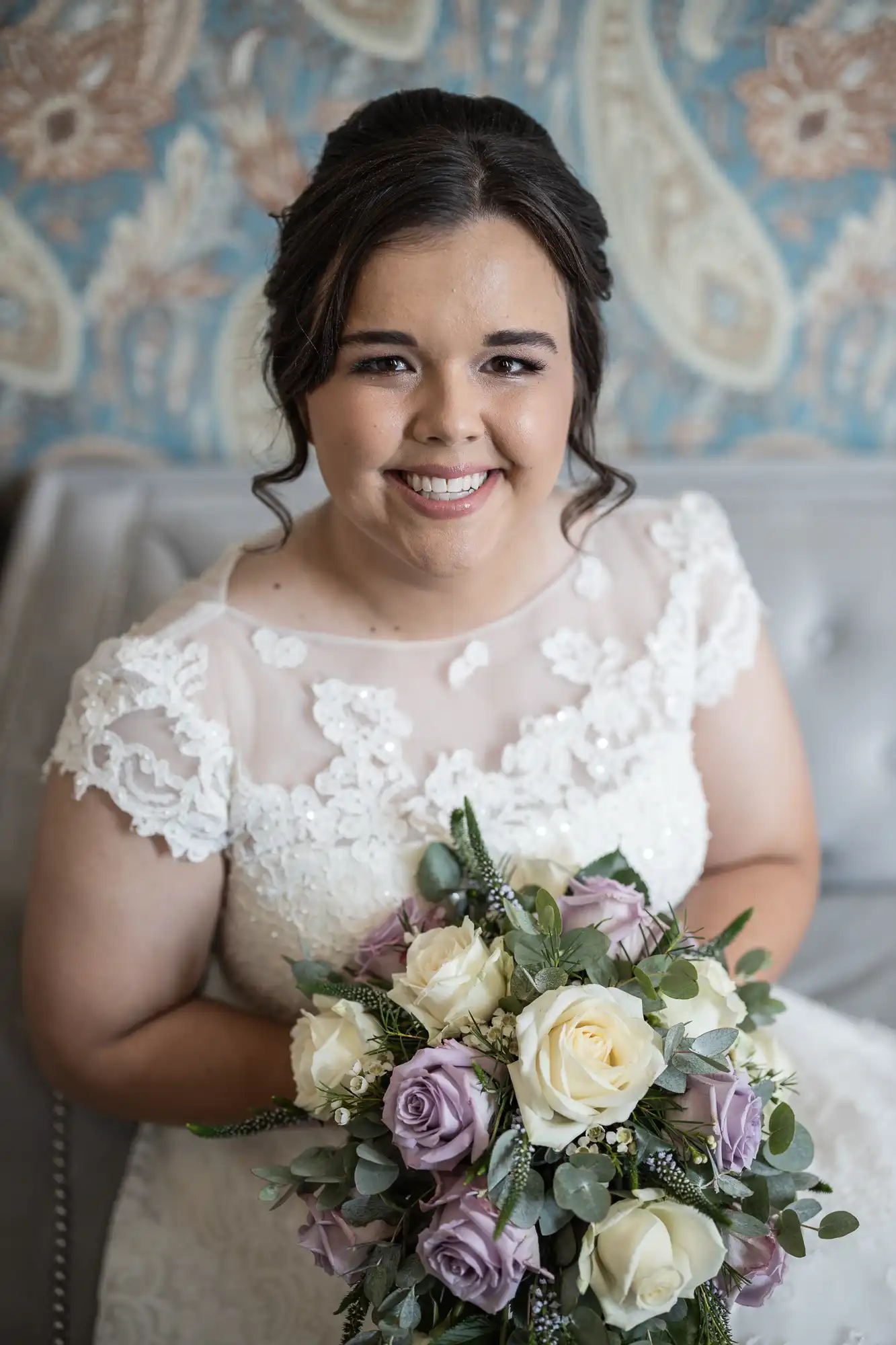 A smiling woman in a white lace dress holds a bouquet of white and light purple roses, sitting on a light-colored sofa with a decorative paisley backdrop.