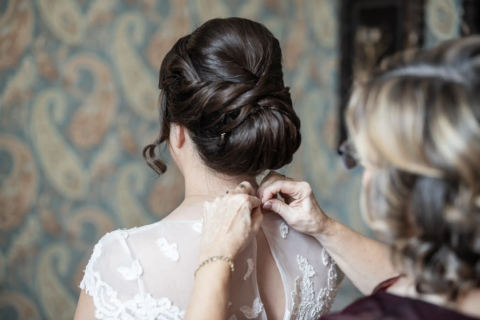 Woman helps bride in a white lace dress button the back of her gown, against a patterned wallpaper background.