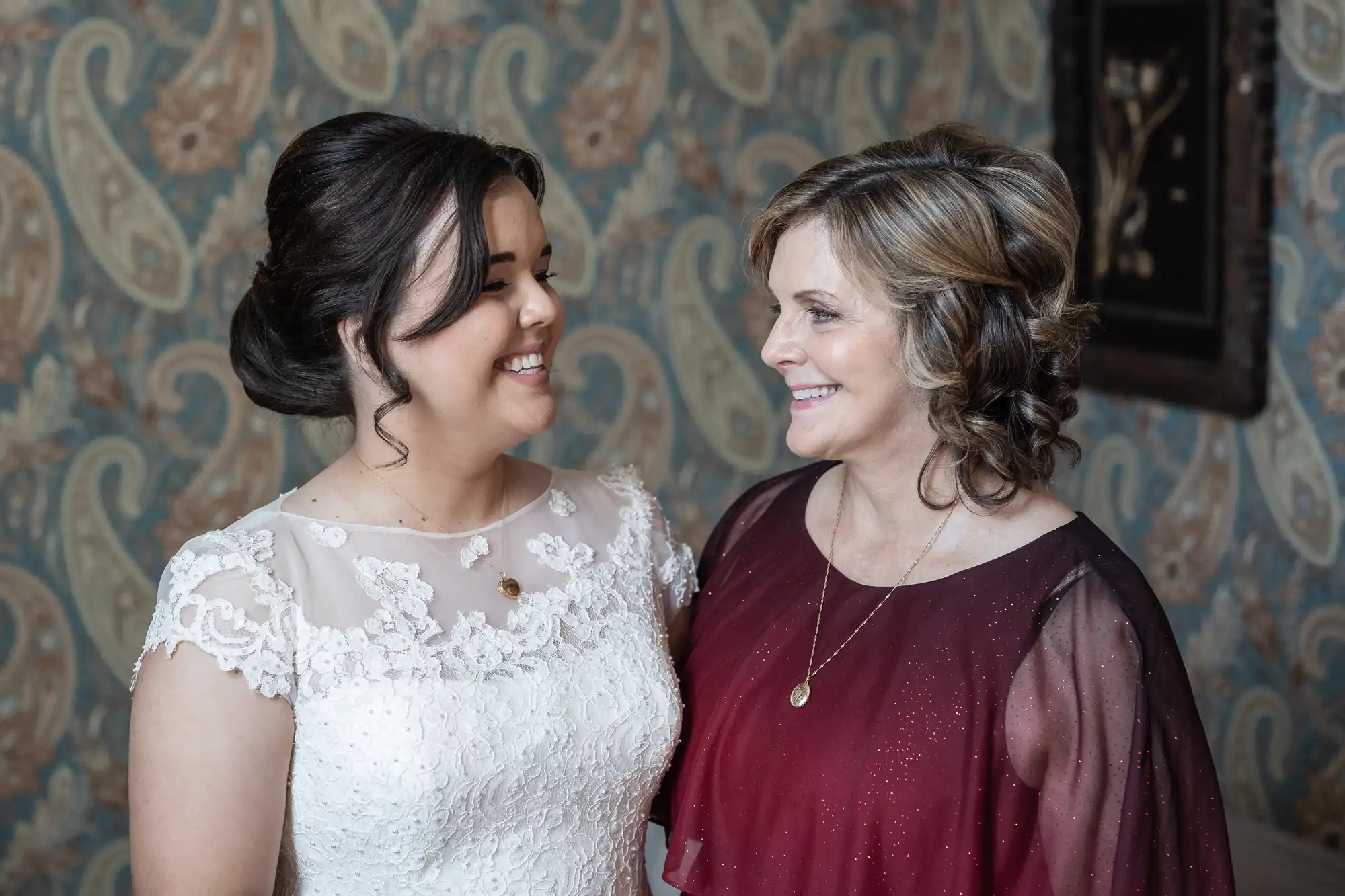 Two women standing side by side, smiling at each other. One is wearing a white lace dress, and the other is in a maroon dress. The wallpaper behind them has a paisley pattern.