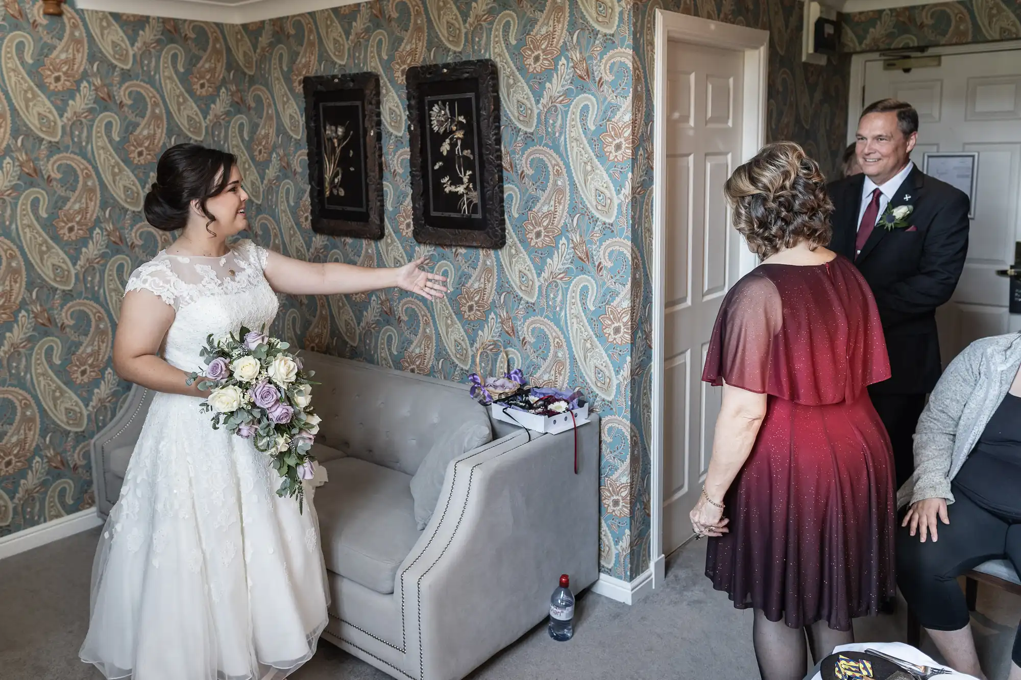 A bride in a white dress extends her arm toward a woman and a man standing in a room with patterned wallpaper. The man is in a suit, and the woman wears a red gradient dress.