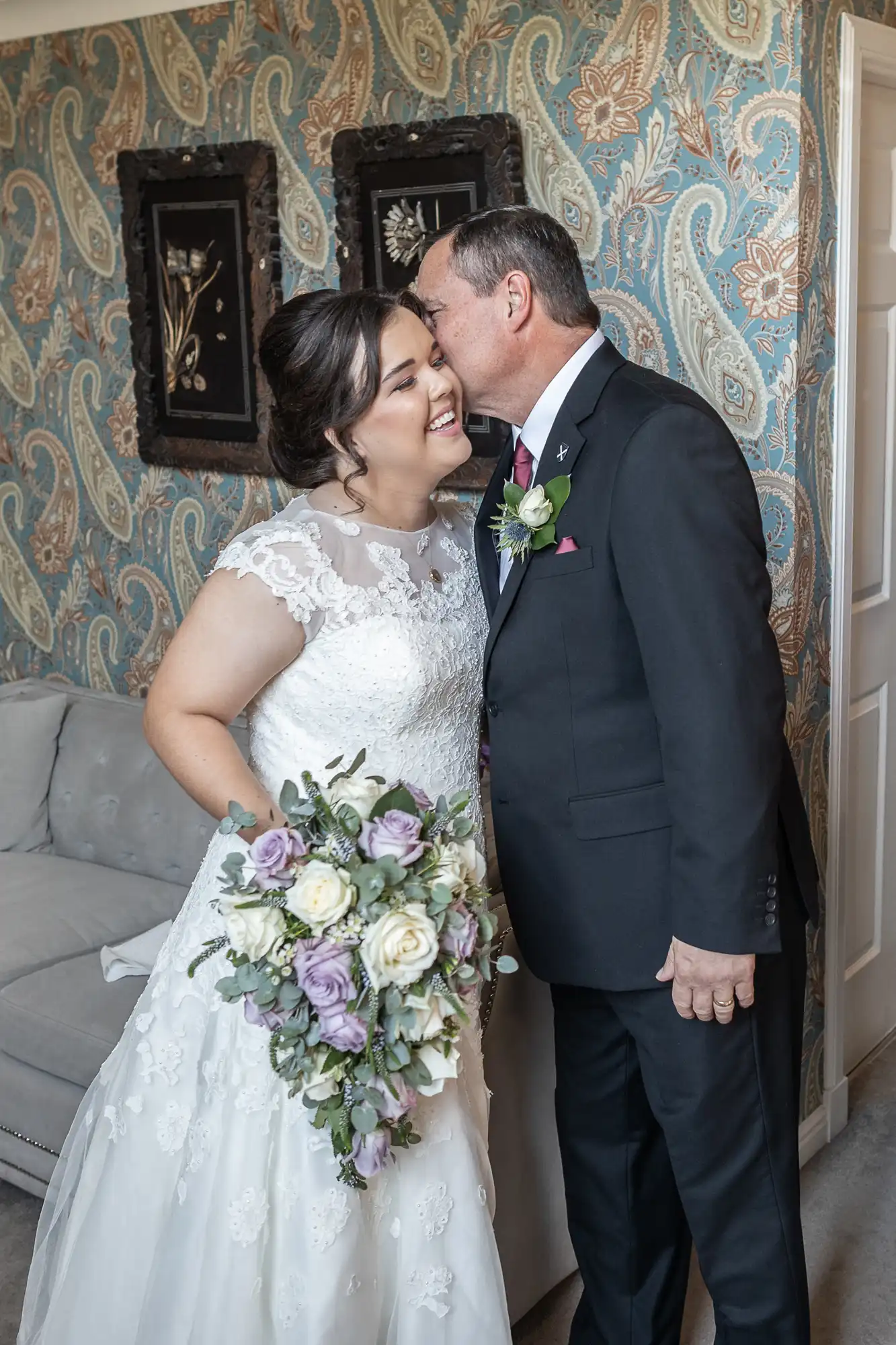 A bride in a white lace wedding dress holding a bouquet of white and purple flowers smiles as an older man in a dark suit and tie whispers in her ear. They are in a room with patterned wallpaper.