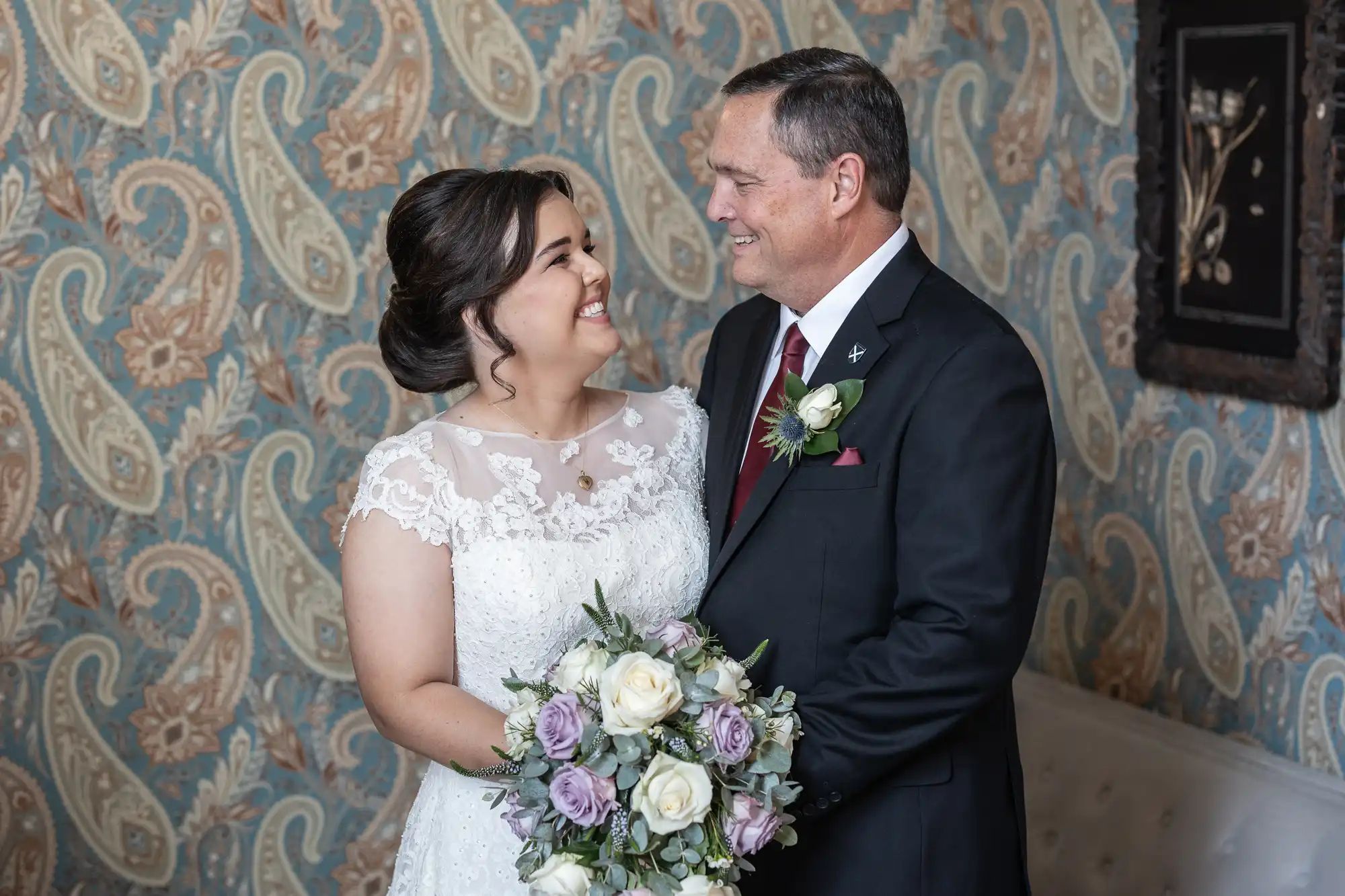 A bride in a white dress and a groom in a black suit with a red tie smile at each other while holding a bouquet of white and purple flowers, standing indoors against a patterned wallpaper.