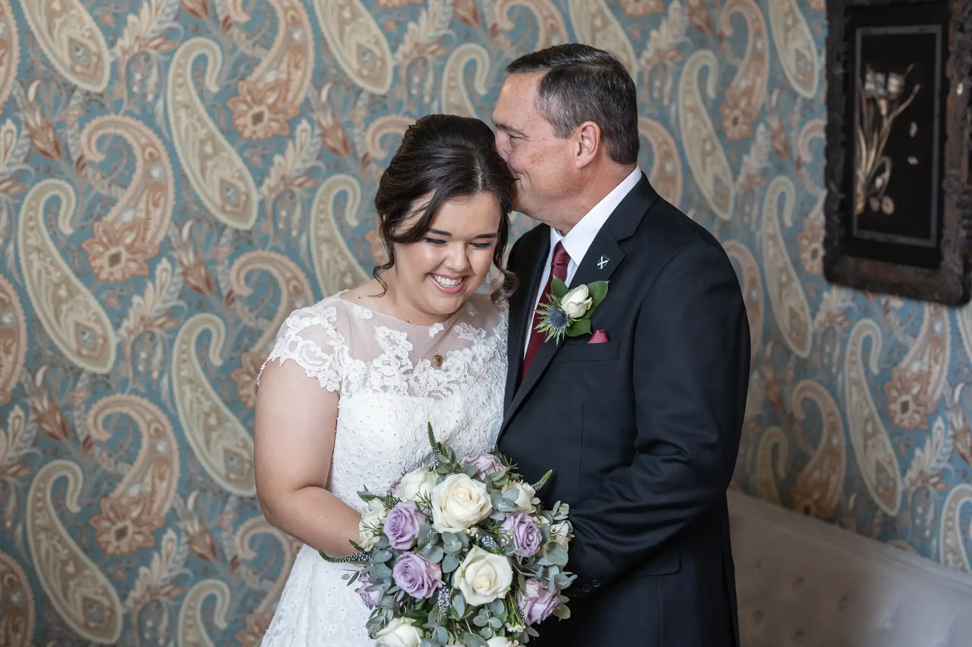 A bride in a white lace dress and holding a bouquet smiles as an older man in a black suit with a red tie kisses her on the forehead in a room with paisley-patterned wallpaper.