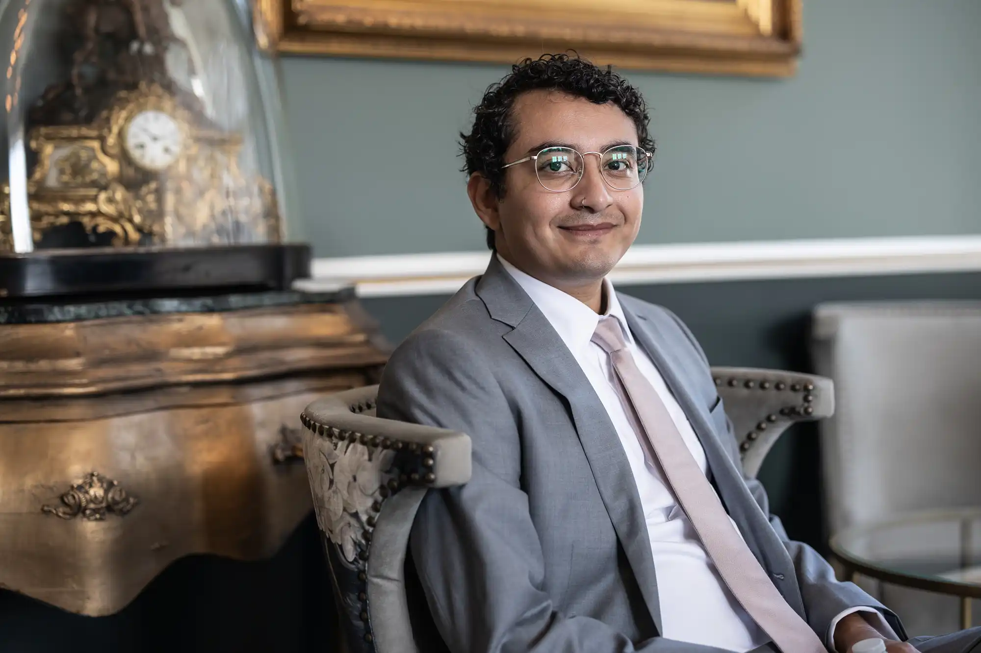 A man in a grey suit and glasses sits on a chair, smiling, with a vintage clock and framed picture in the background.