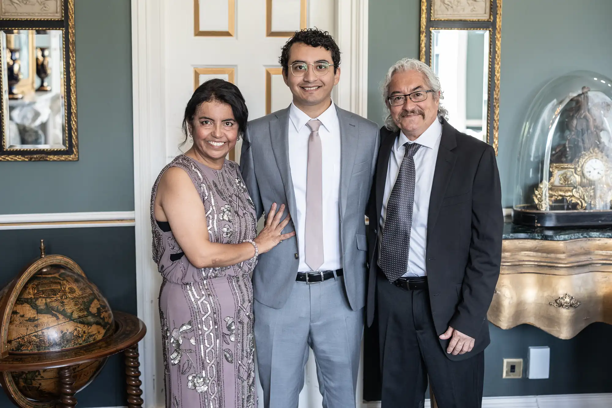 Three people stand closely together, smiling at the camera. The woman on the left wears a patterned dress, the man in the center wears a gray suit, and the man on the right wears a dark suit and tie.