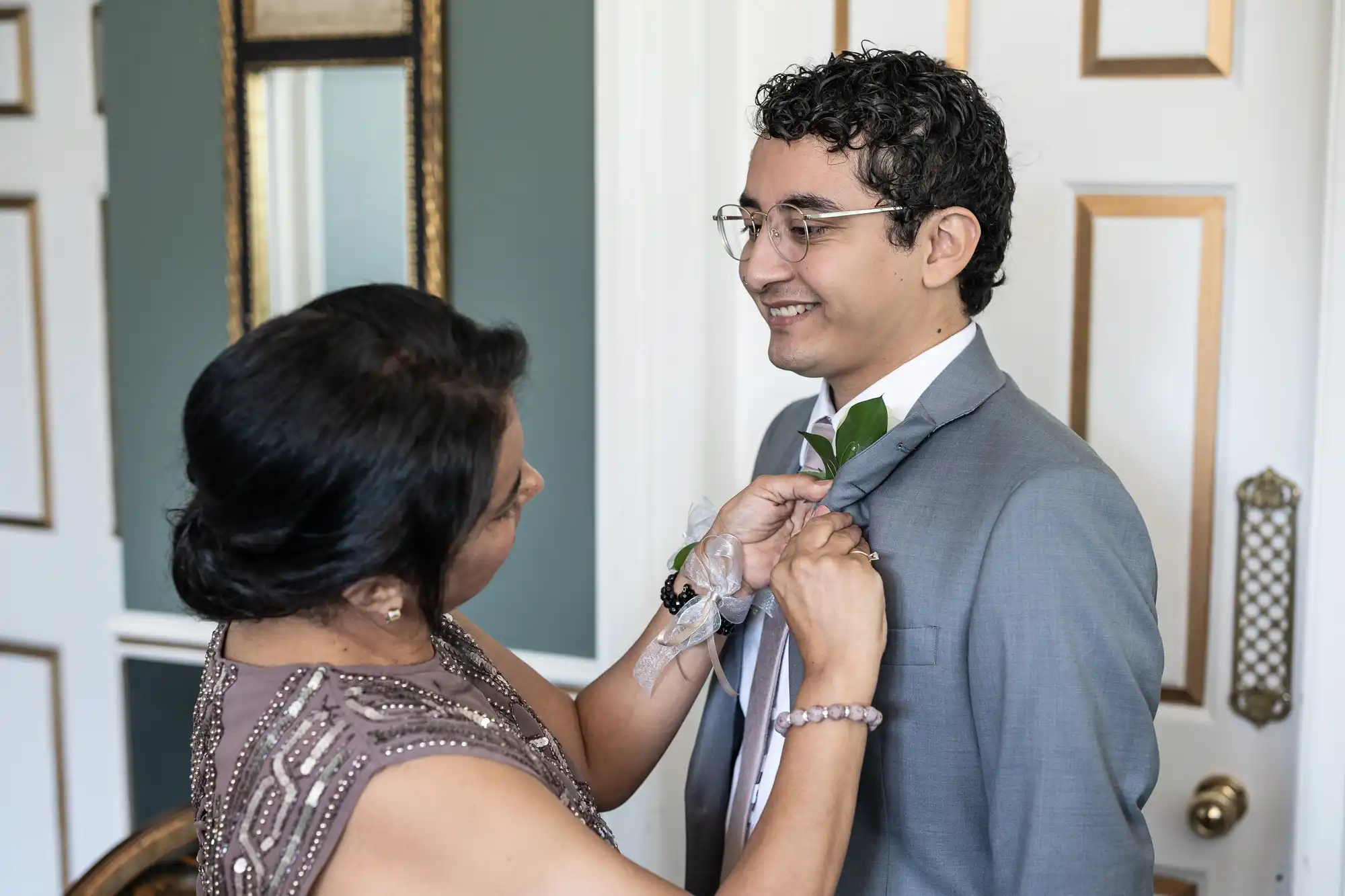 A woman adjusts a boutonniere on a smiling man wearing a gray suit in a room with ornate décor.