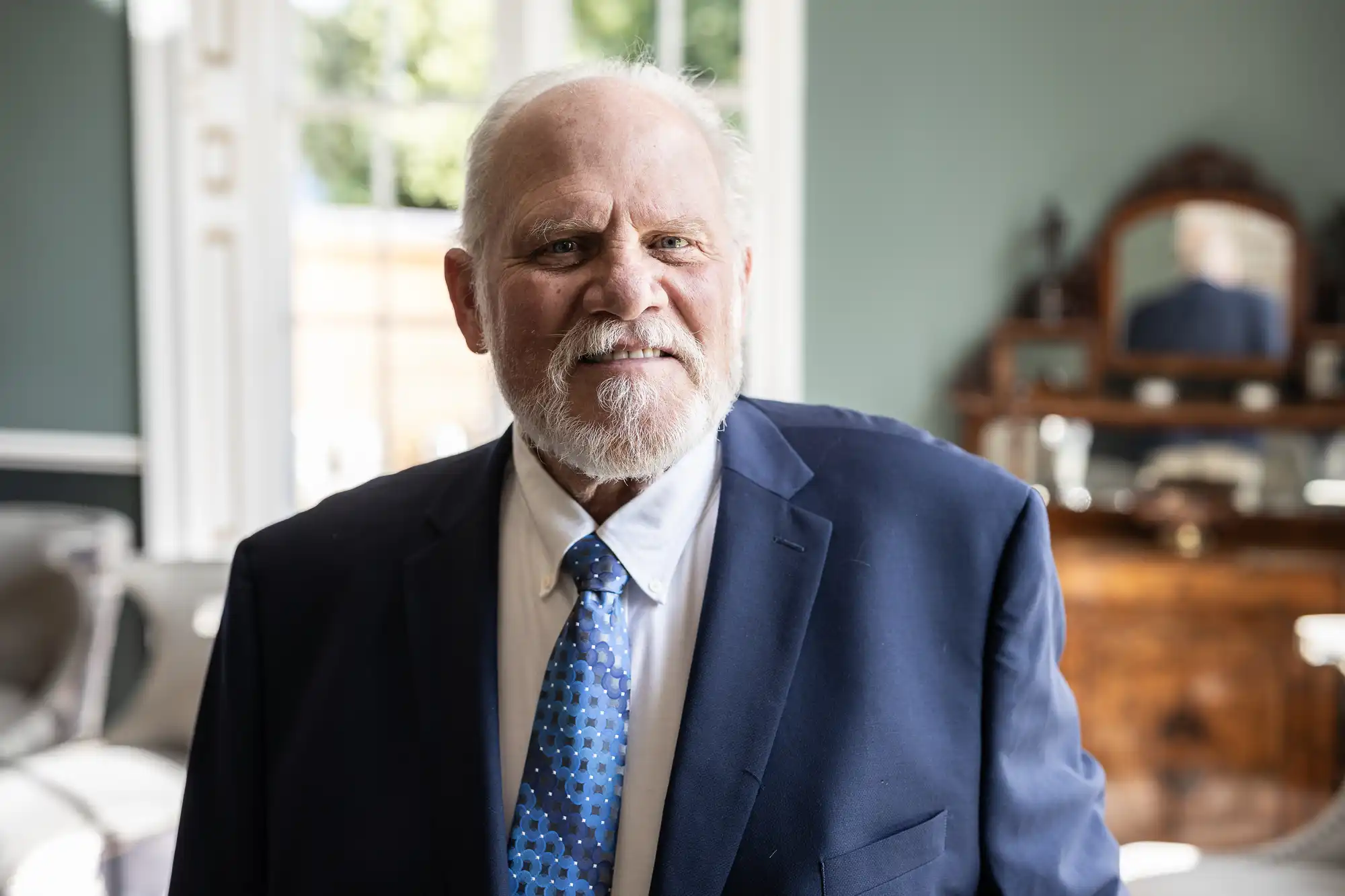 An older man with a white beard wearing a blue suit and tie stands indoors in front of a dresser with a mirror.