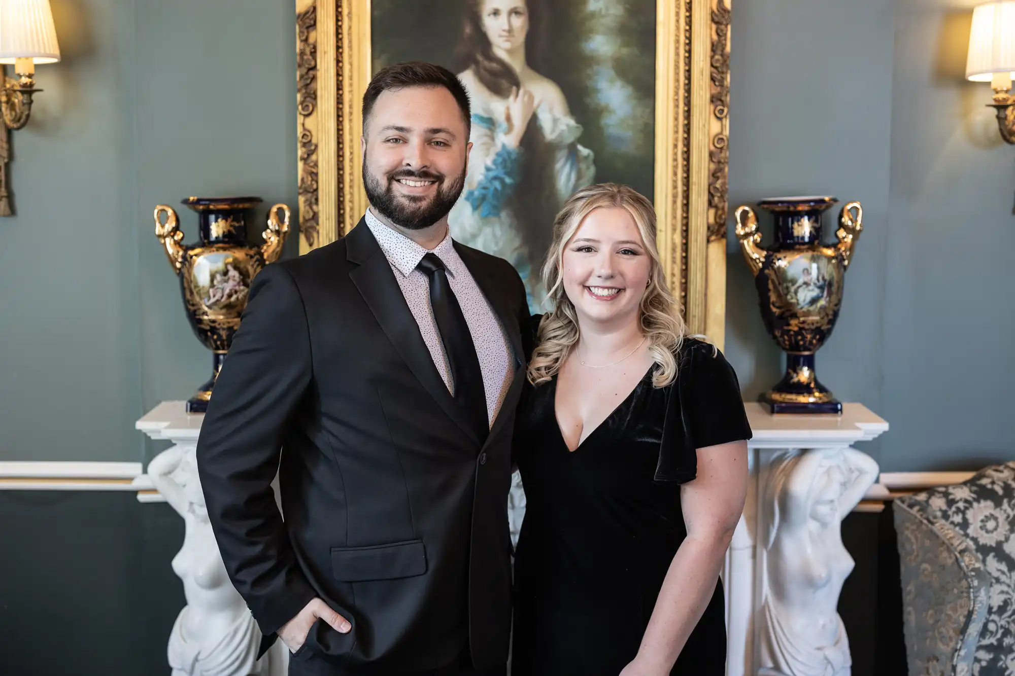 A man and a woman, both dressed formally, stand smiling in front of a framed portrait and two blue vases.