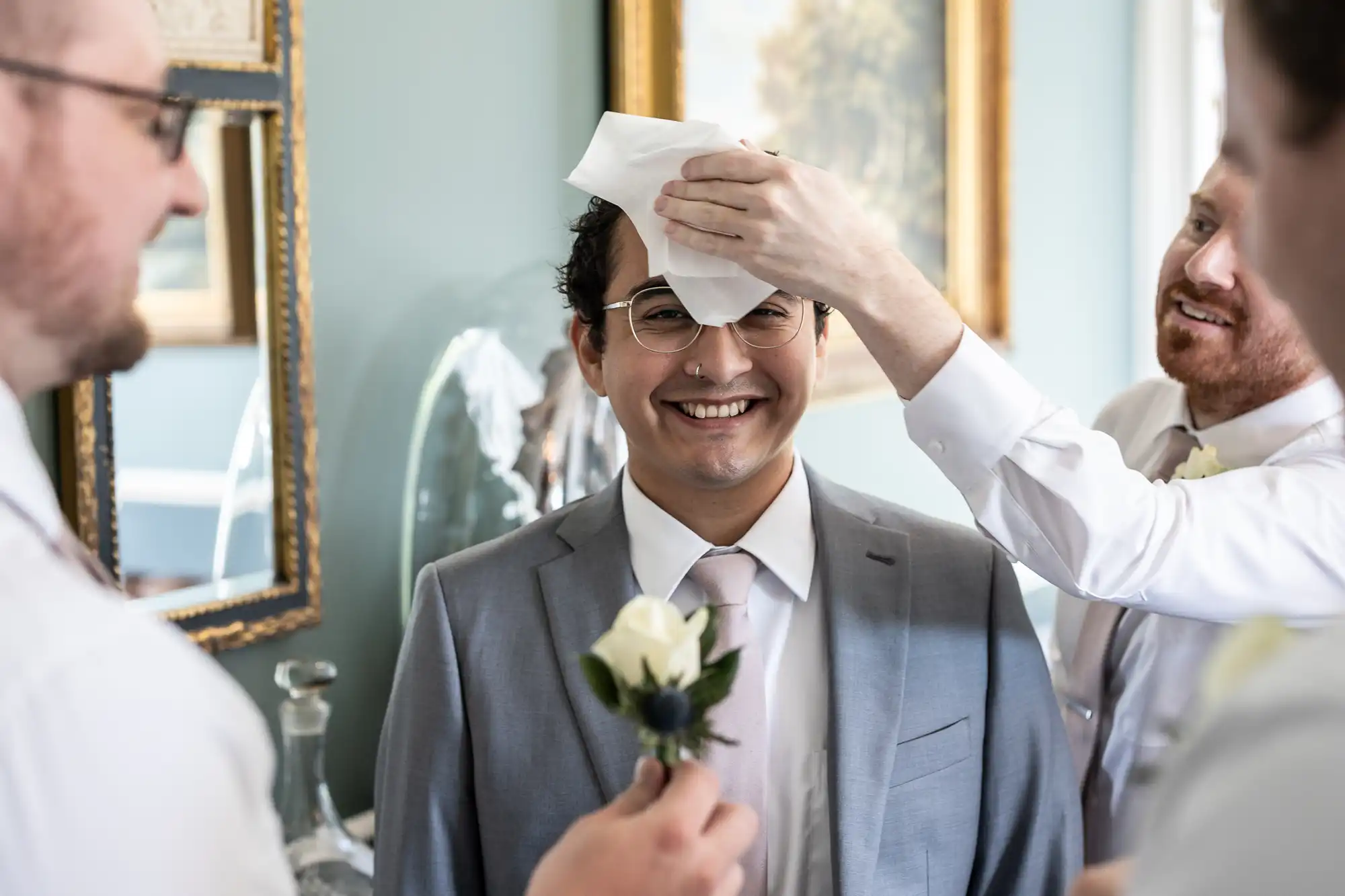 Groom having his forehead dabbed with a tissue by one groomsman while another holds a boutonnière in front of him; all smiling in a softly lit room.