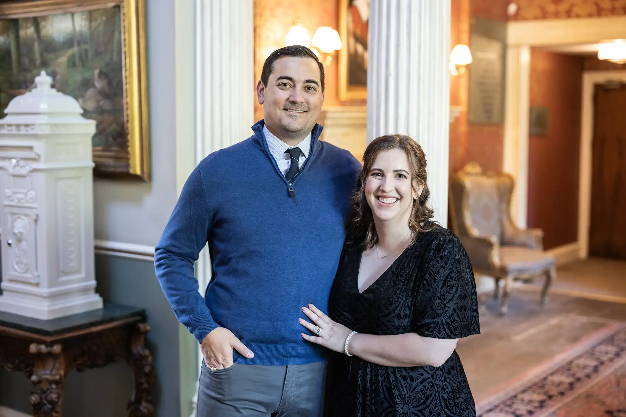 A man in a blue sweater and gray pants stands beside a woman in a black dress, smiling in an elegantly decorated room with framed artworks and Victorian-style furniture.