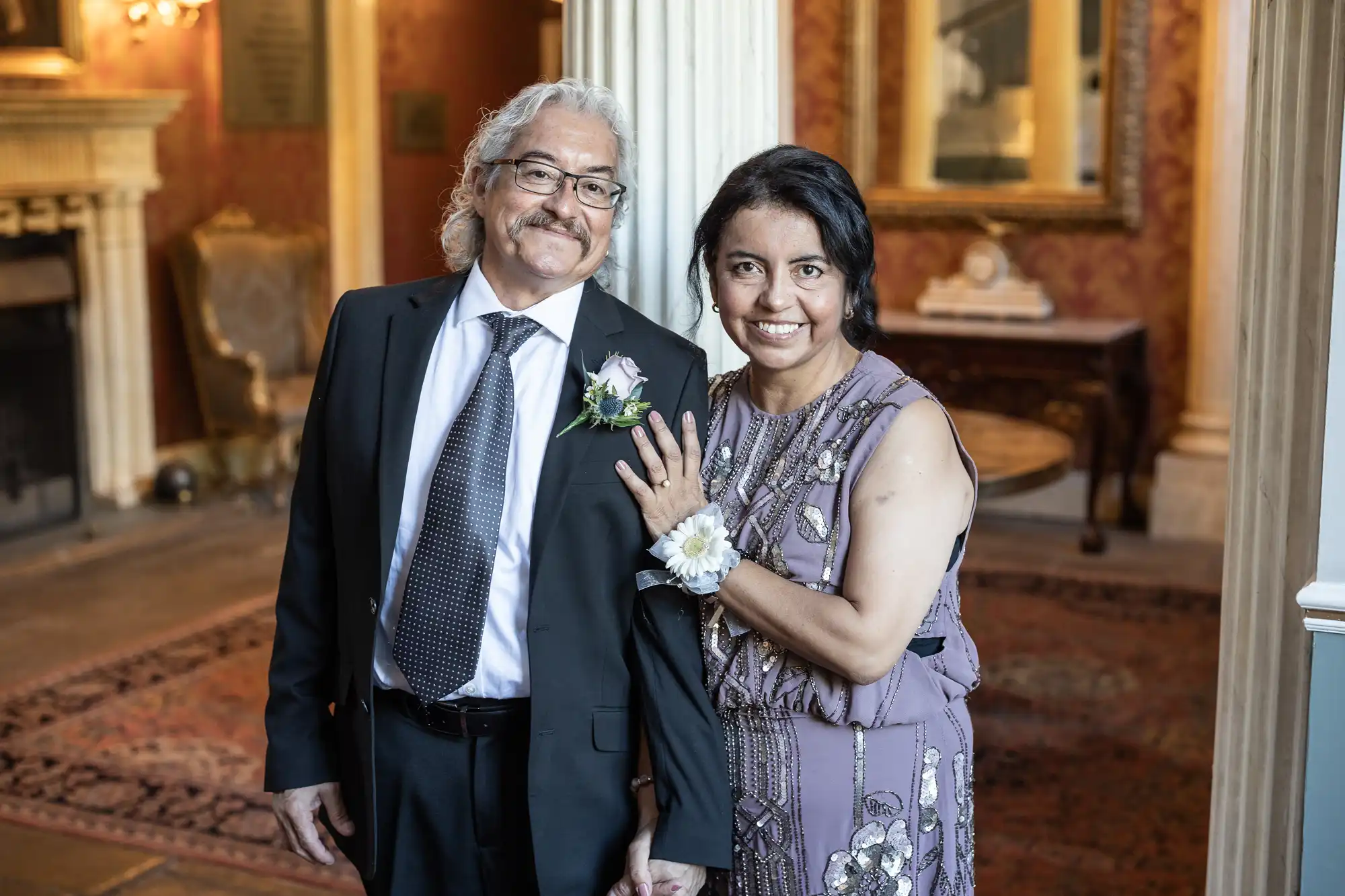 A couple dressed formally stands indoors smiling at the camera. The woman has her hand on the man's chest, wearing a corsage. Both are in front of an ornate background with columns and framed artwork.