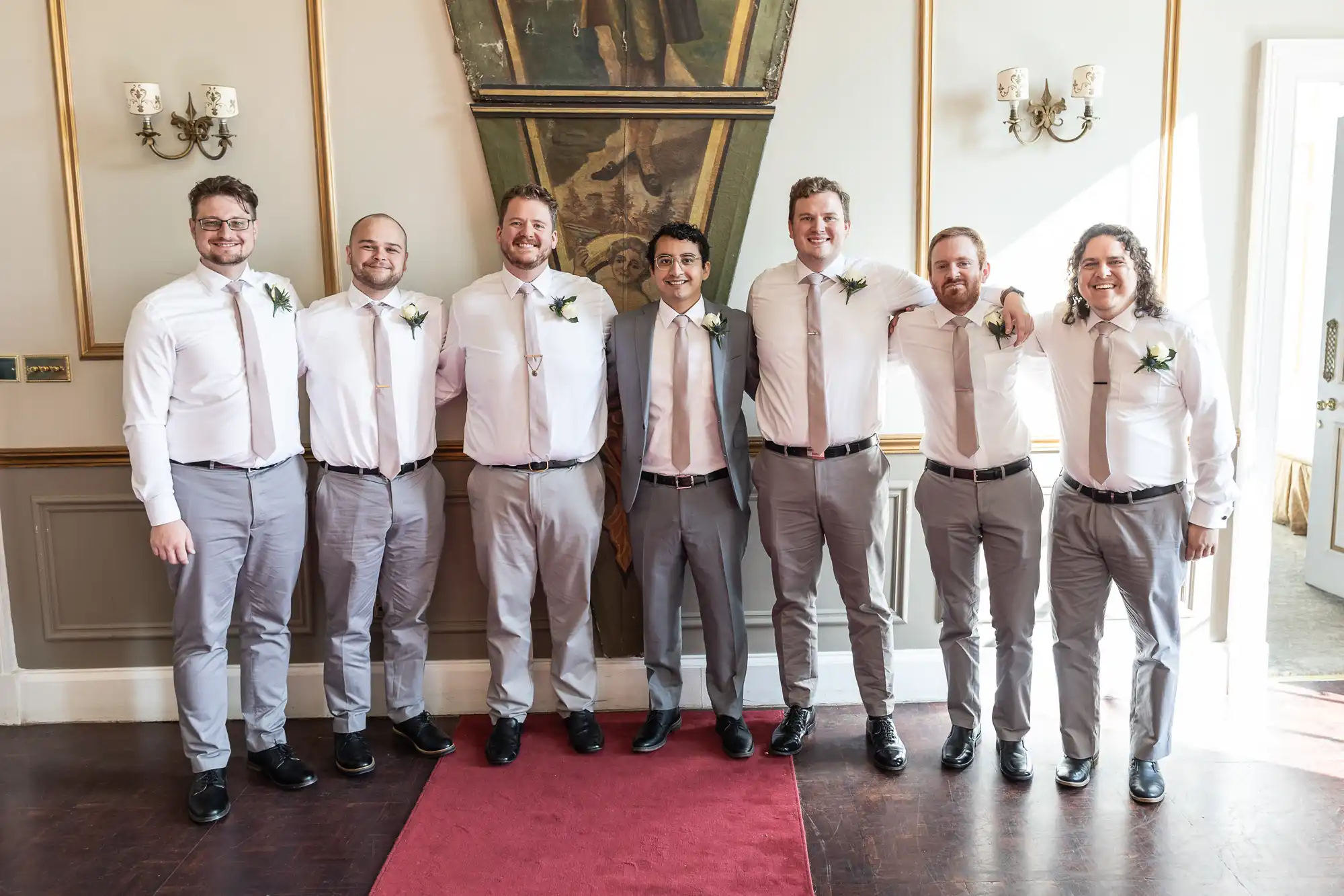 A group of seven men dressed in light-colored shirts and pants, some with ties, stand on a red carpet in an ornate room, posing for a photo. All are smiling and wearing boutonnieres.