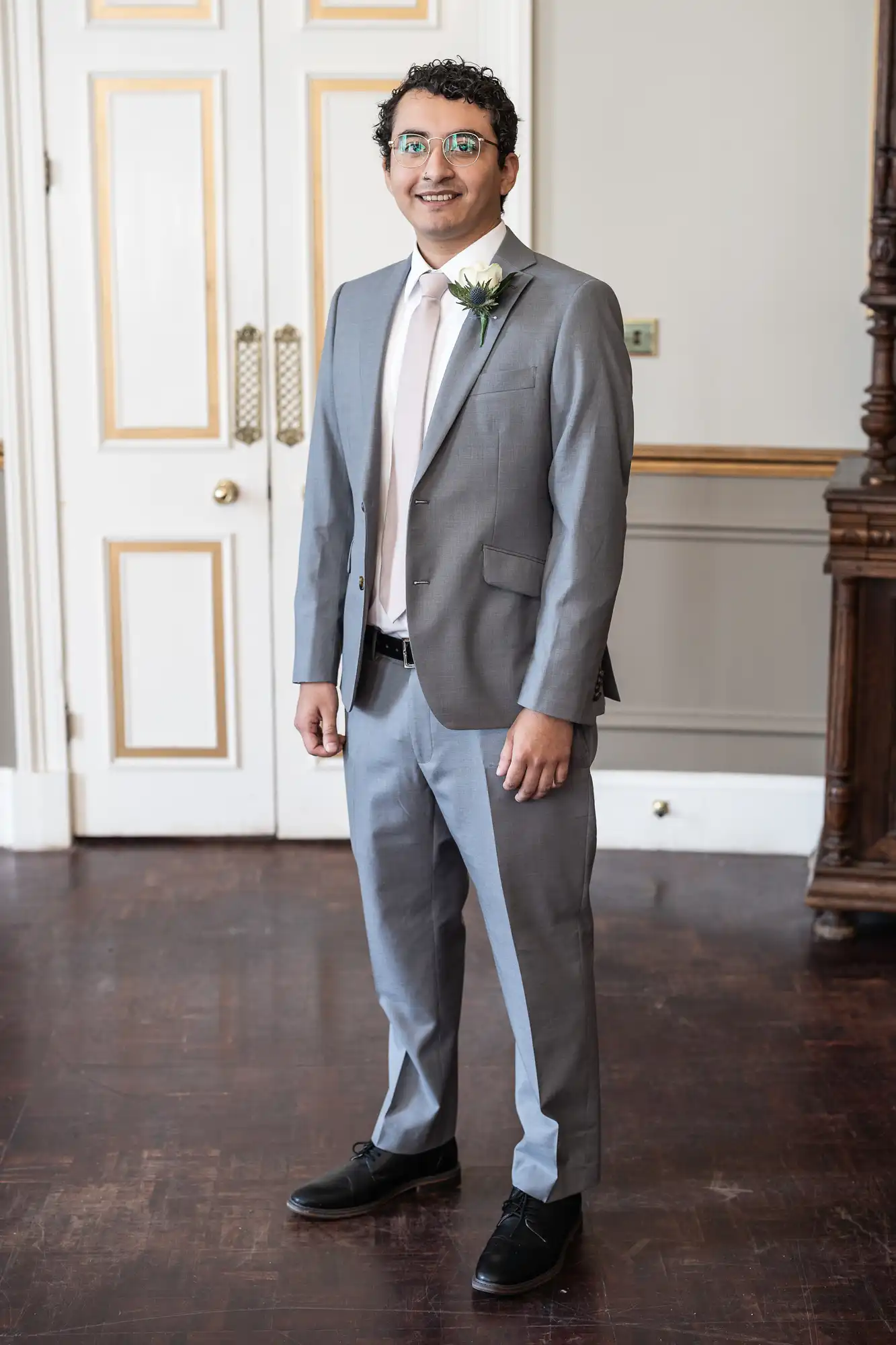 A man in a grey suit with a white shirt and light pink tie stands indoors in front of double doors. He is wearing glasses and has a white flower pinned to his lapel.